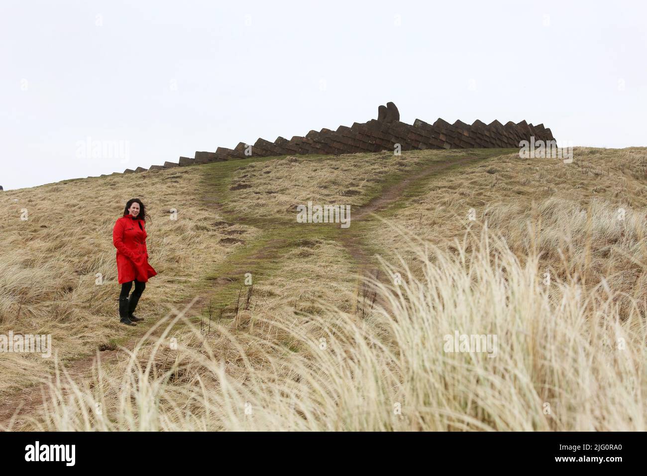 Donne cauasiche bianche di età media che indossano un cappotto rosso trenchcoat , wellingtons neri da soli su spiaggia bagnata e ventosa come le onde si rompono, isolate, premurose, enigmatiche con spazio per la copia e il testo Foto Stock