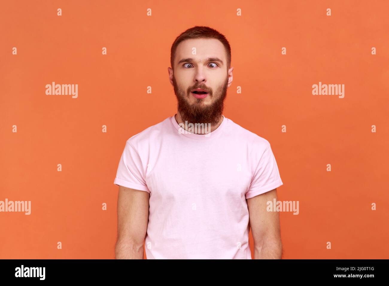 Ritratto di uomo bearded che osserva gli occhi incrociati con il sorriso stupido, ingannando intorno, facendo il viso sciocco, espressione comica senza cervello, indossando la T-shirt rosa. Studio interno girato isolato su sfondo arancione. Foto Stock