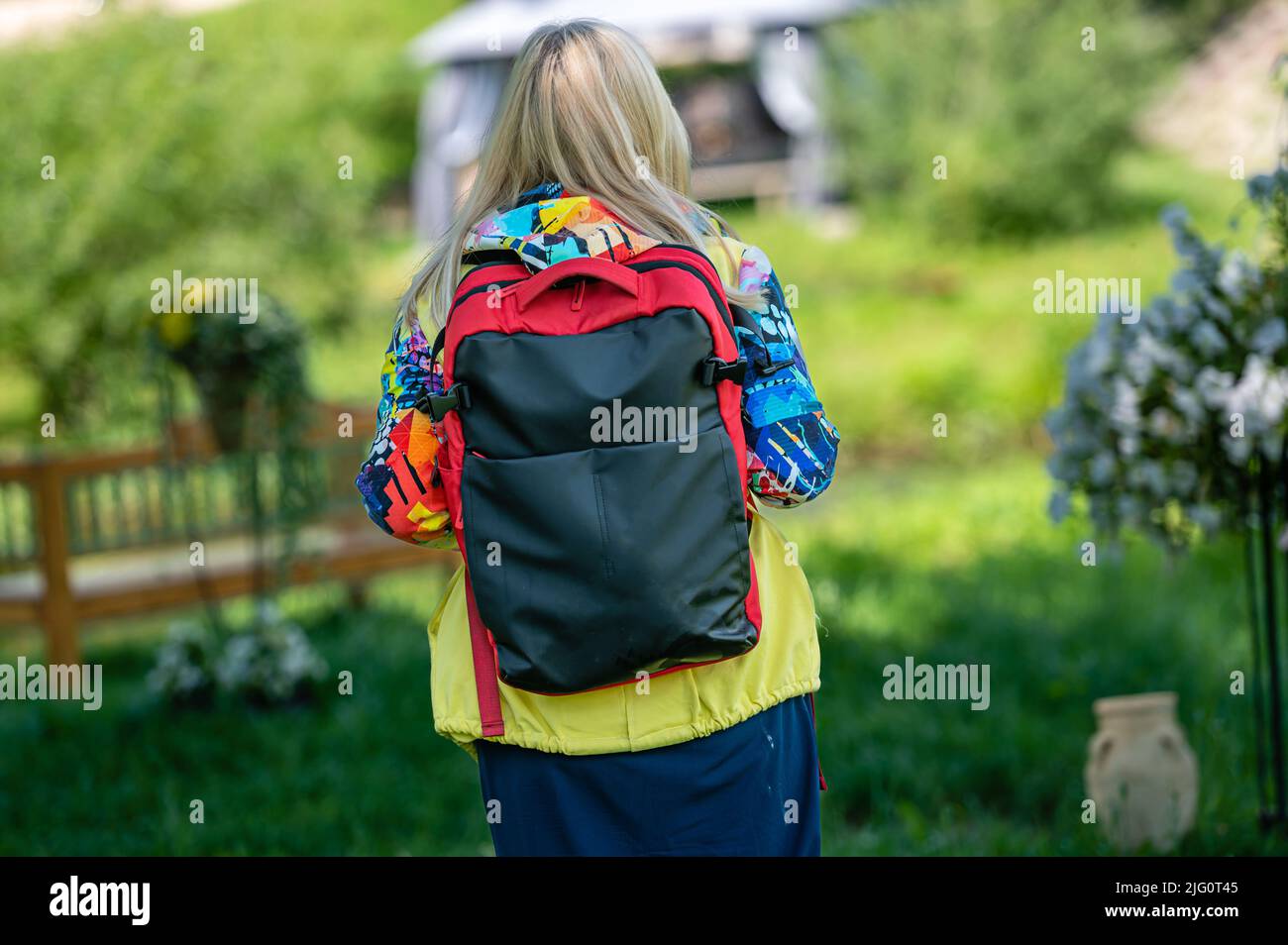 donna anziana bionda in abiti luminosi con uno zaino nel parco in una giornata di sole, vista posteriore Foto Stock