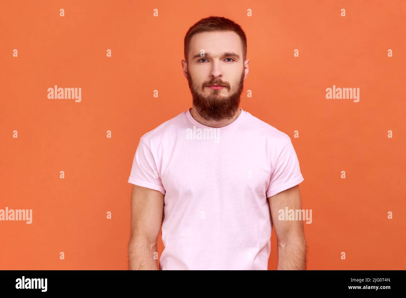 Ritratto di uomo bearded sicuro di sé, guardando la macchina fotografica con espressione seria, ragazzo unsmiling determinato, indossando la T-shirt rosa. Studio interno girato isolato su sfondo arancione. Foto Stock