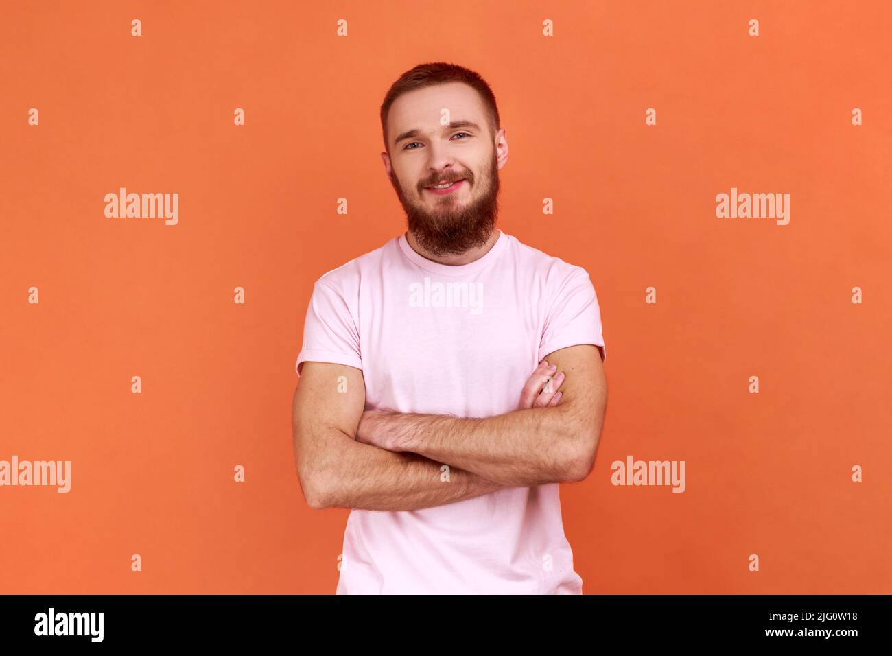 Ritratto di uomo barbuto in piedi con le braccia incrociate, guardando la macchina fotografica, esprimendo emozioni positive, essendo di buon umore, indossando una T-shirt rosa. Studio interno girato isolato su sfondo arancione. Foto Stock