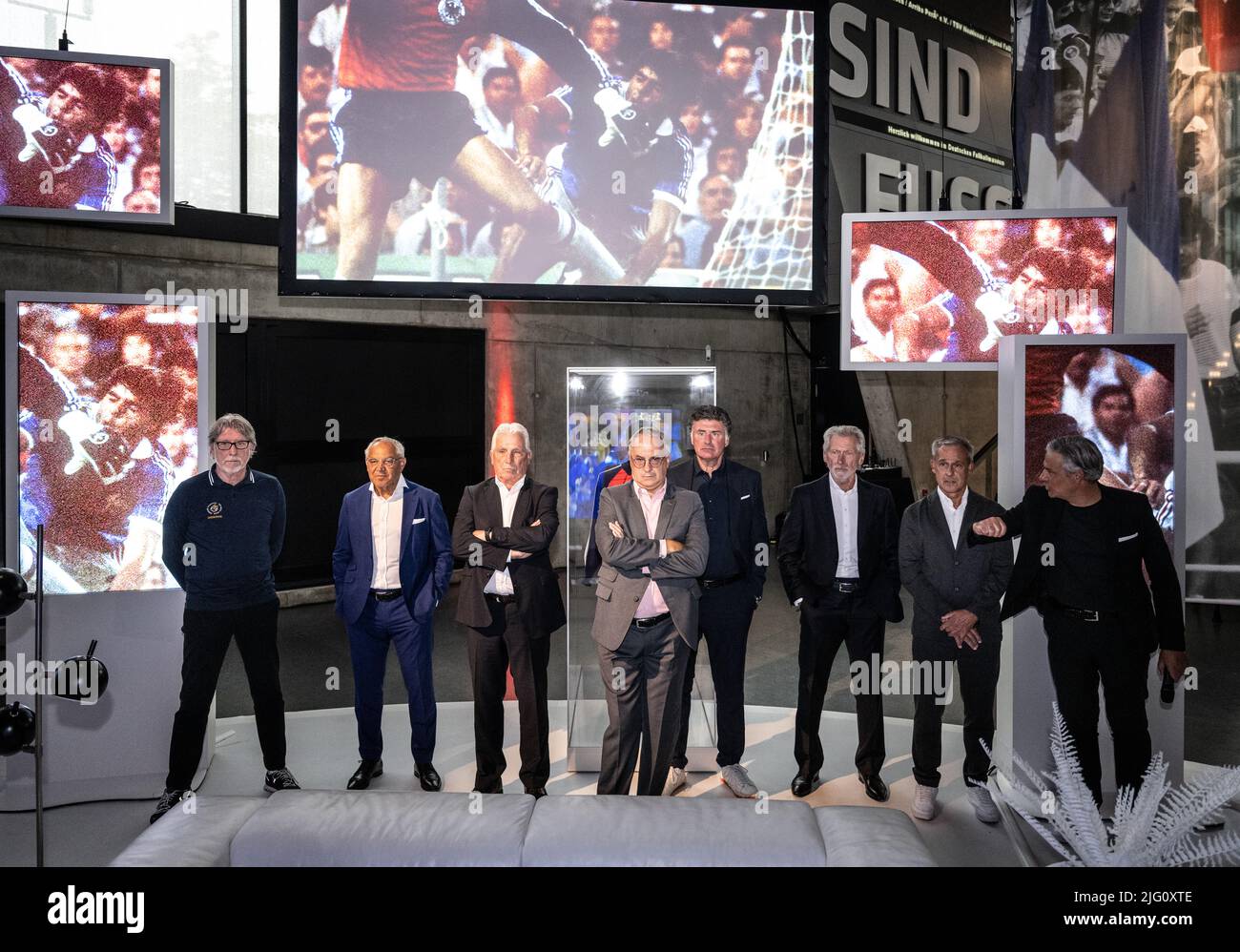 06 luglio 2022, Renania settentrionale-Vestfalia, Dortmund: Ex calciatori nazionali toni Schumacher, Felix Magath, Klaus Fischer, Uli Stielike, Wilfried Hannes, Paul Breitner e Pierre Littbarski, nonché Manuel Neukirchner, direttore del Museo tedesco del calcio, (da sinistra) guardano estratti della semifinale della Coppa del mondo 1982 contro la Francia su grandi monitor nella mostra speciale per il 40th anniversario della Notte di Siviglia al Museo tedesco del calcio, Su cui si sovrappone un fallo di Schumacher sul giocatore francese Patrick Battiston. Nel 40th anniversario della Notte di Siviglia, il Foto Stock
