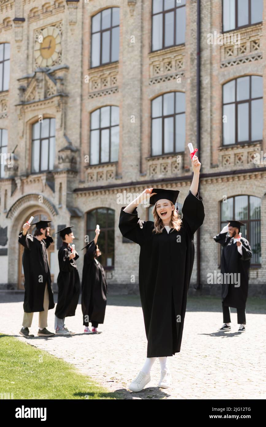 Bachelor eccitato che tiene il diploma vicino a blurred gli amici multietnici e l'università all'aperto Foto Stock