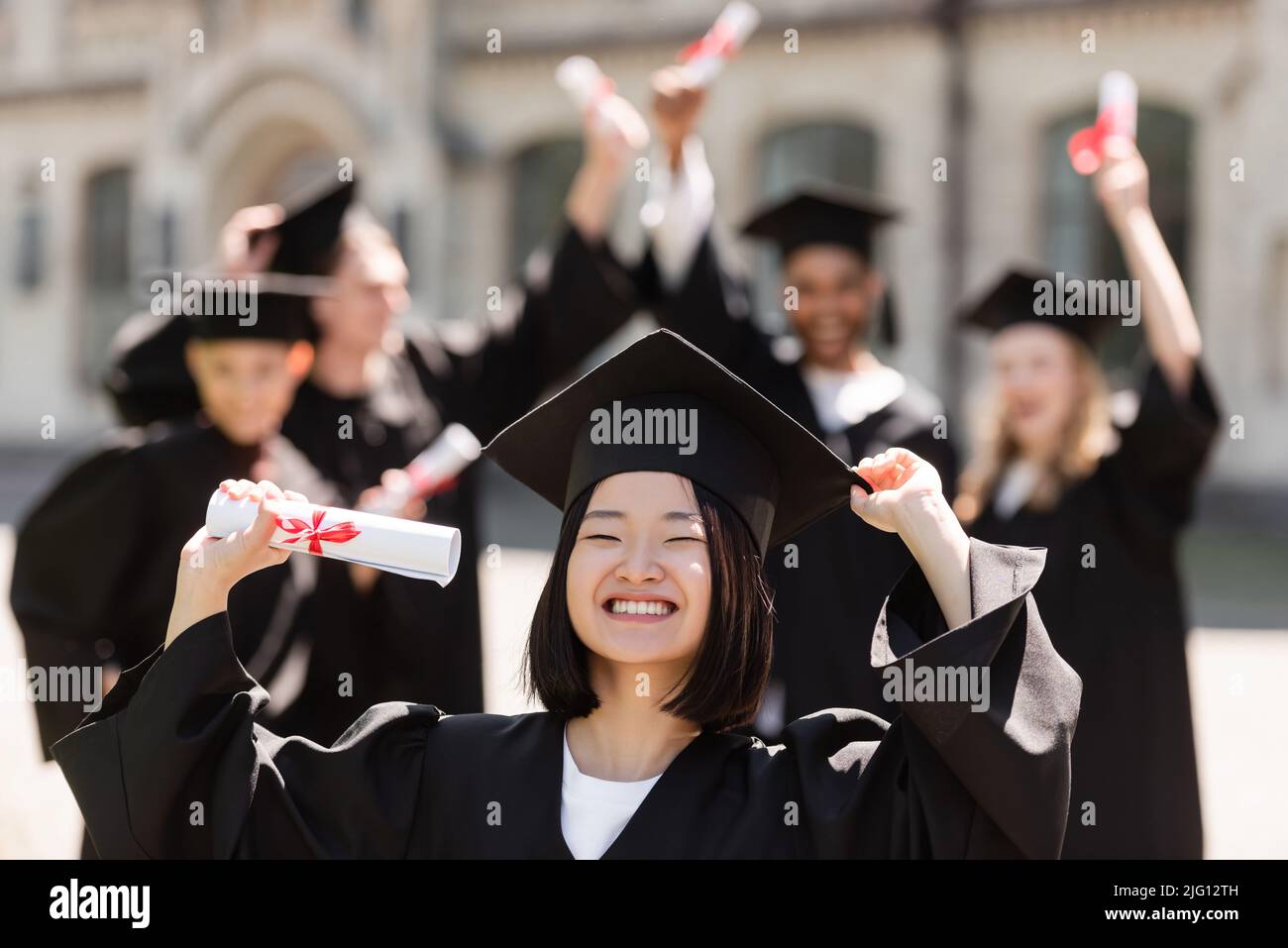 Sorridente bachelor asiatico in berretto e abito che tiene il diploma vicino a blurred amici all'aperto Foto Stock