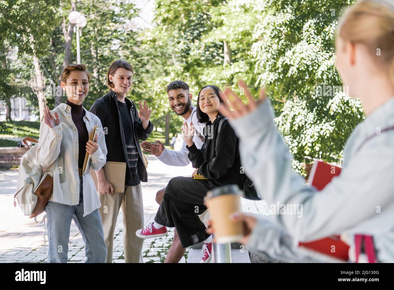 Sorridenti studenti multietnici che agitavano le mani a un amico sfocato nel parco Foto Stock