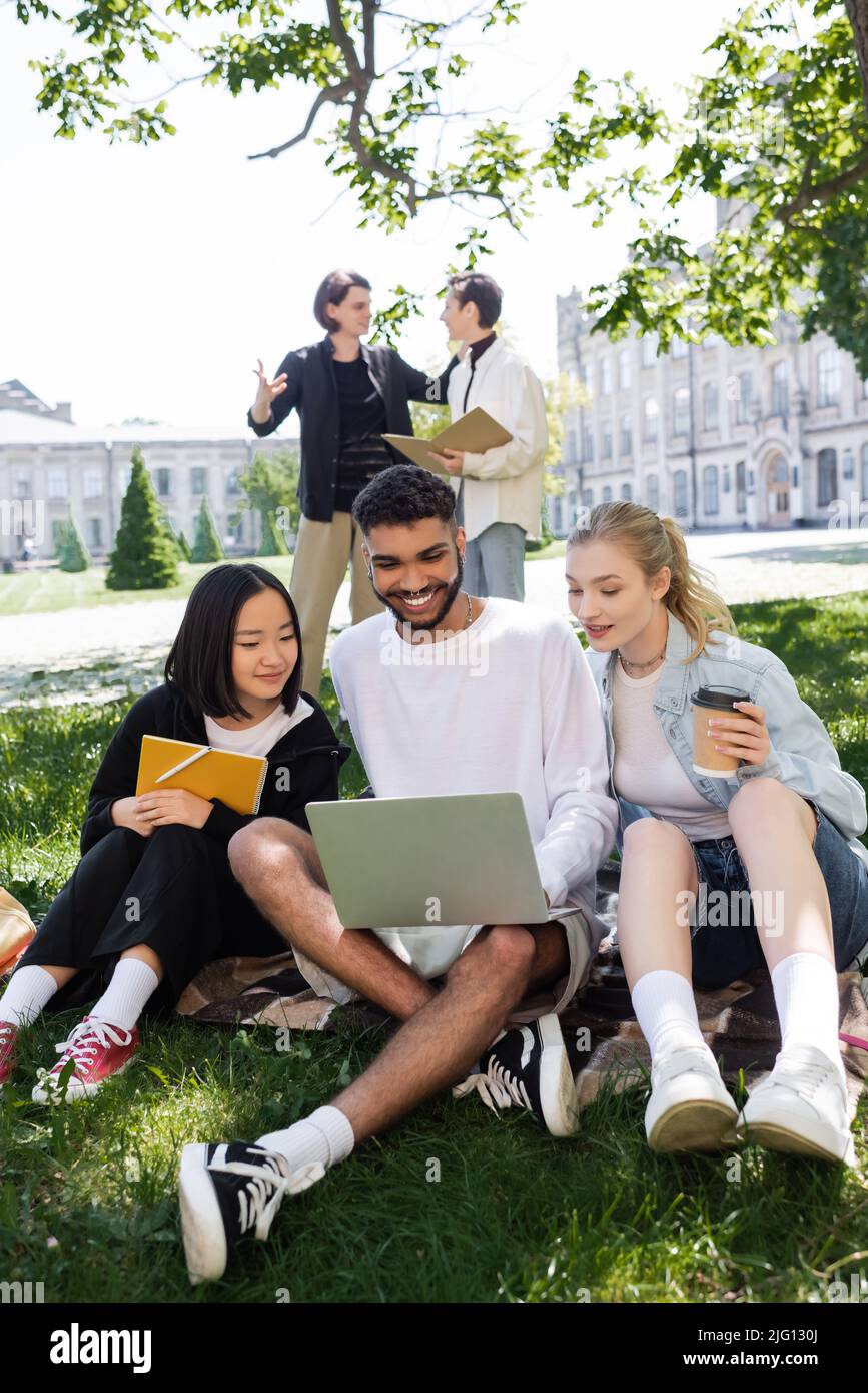Studente afroamericano che usa un computer portatile vicino ad amici multietnici con copia del libro e caffè in erba nel parco Foto Stock