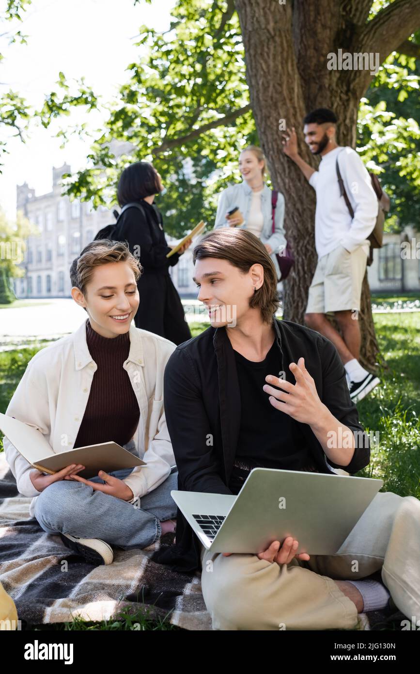 Studente con computer portatile che parla vicino ad un amico con notebook su coperta nel parco Foto Stock
