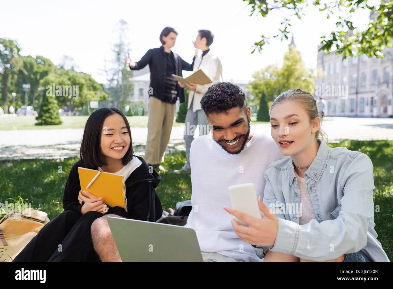 Lo studente tiene smartphone vicino a sorridenti studenti multiculturali con notebook e notebook in erba nel parco Foto Stock