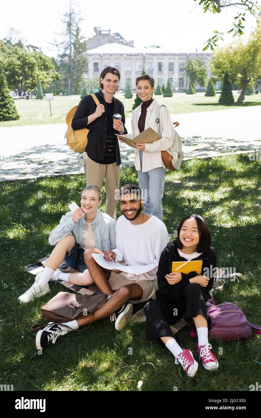 Allegri studenti multietnici con taccuini e caffè guardando la macchina fotografica sul prato nel parco Foto Stock