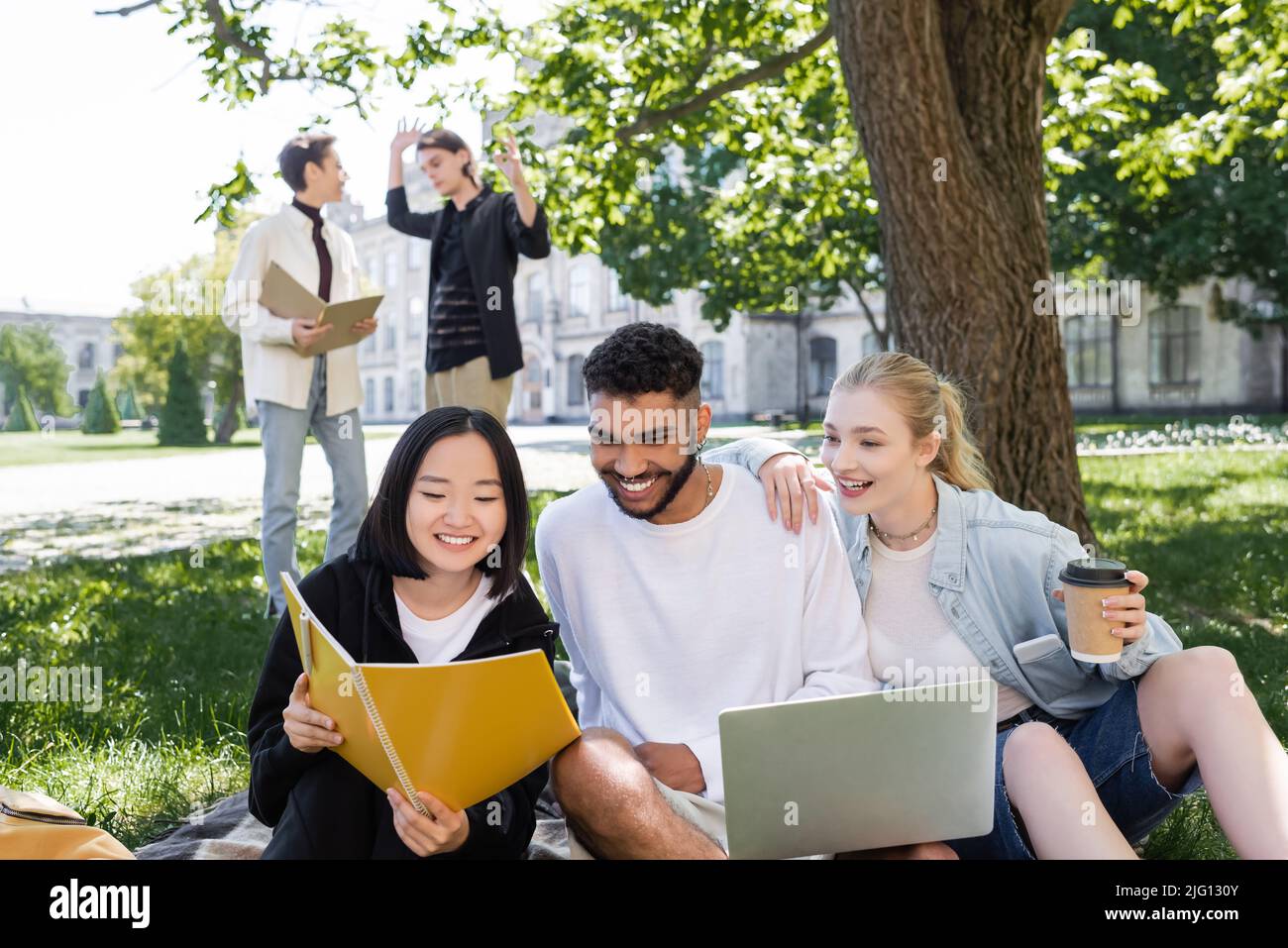 Studente asiatico che tiene notebook vicino agli amici con computer portatile e caffè nel parco Foto Stock