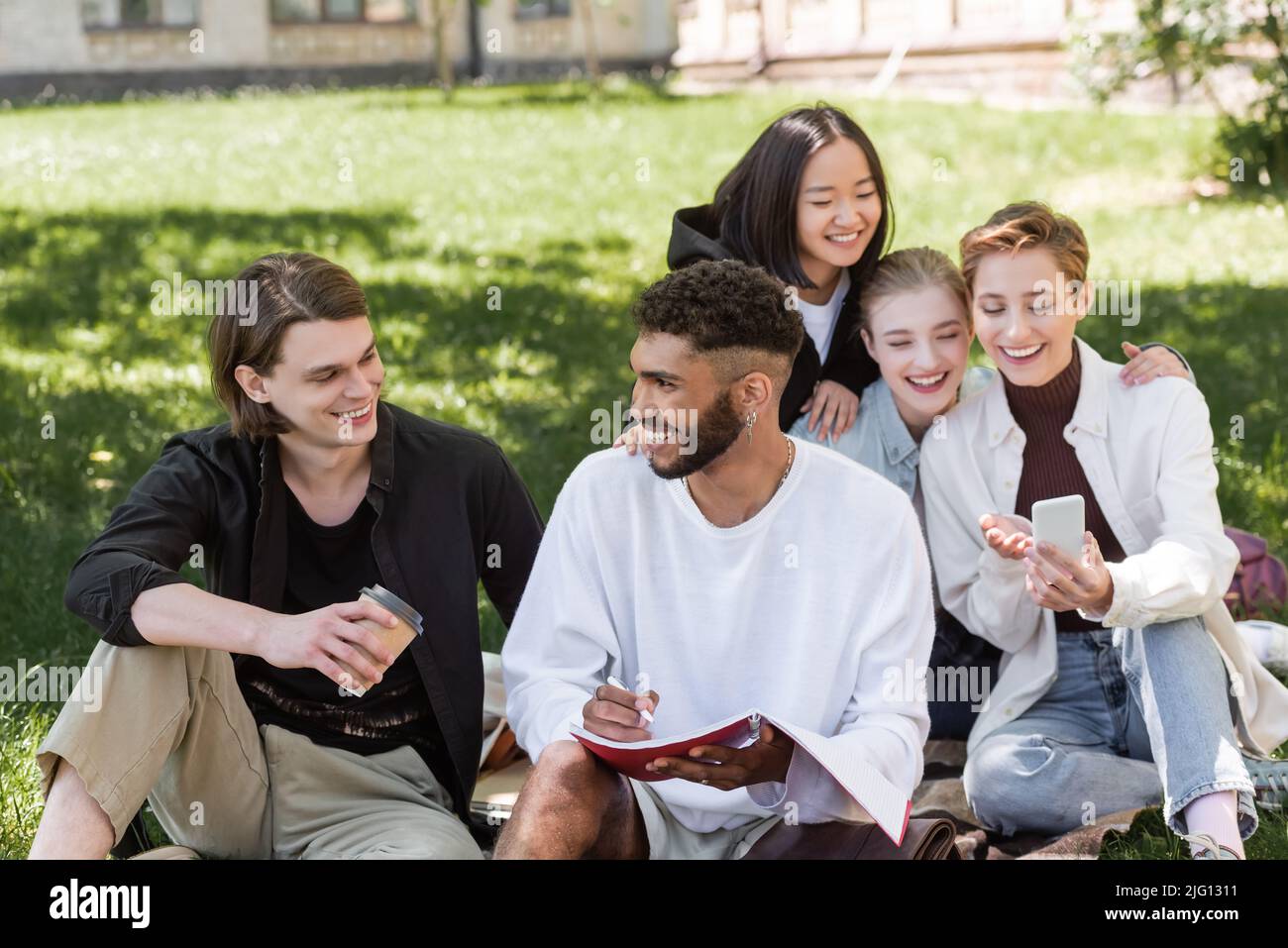 Sorridenti studenti interrazziali che parlano vicino a amici sfocati utilizzando smartphone sul prato nel parco Foto Stock