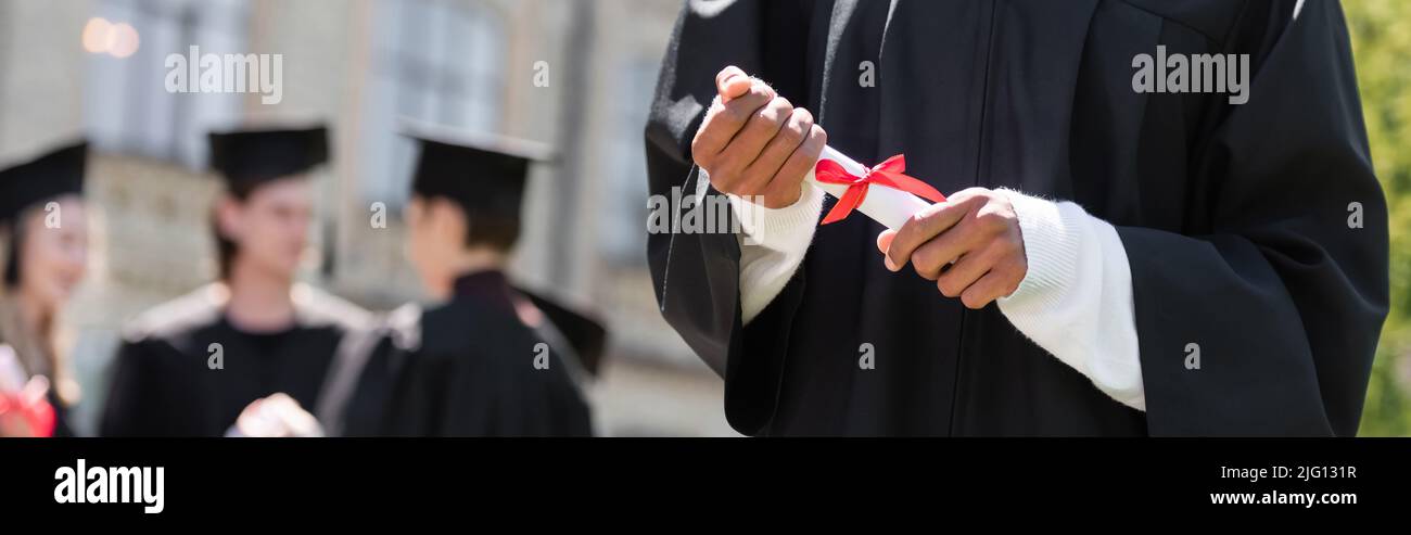Vista croppata del bachelor afroamericano che tiene il diploma vicino agli amici sfocati nel parco, bandiera Foto Stock
