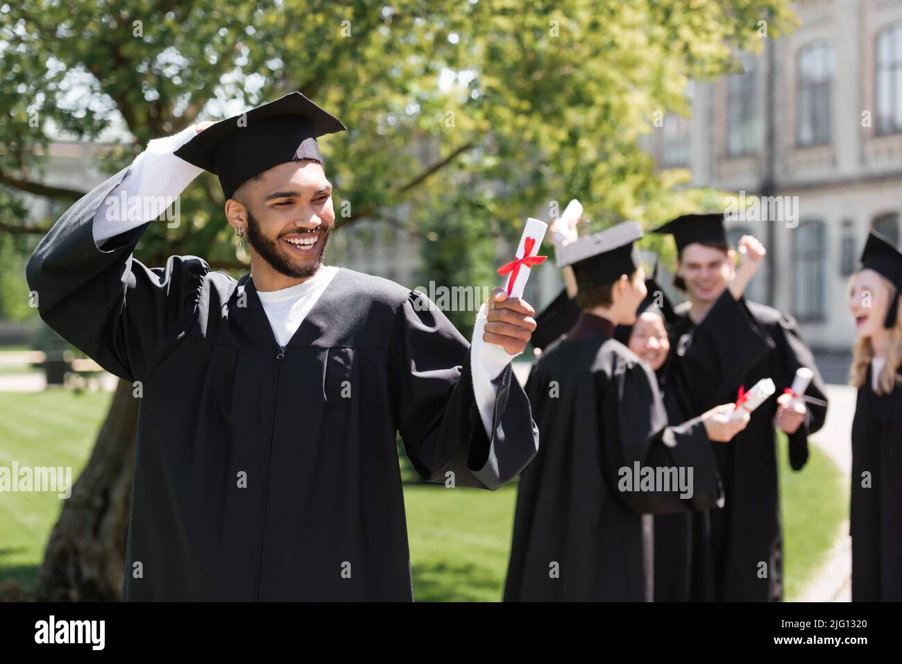 Cheerful african american bachelor guardando il diploma vicino blurred amici nel parco Foto Stock