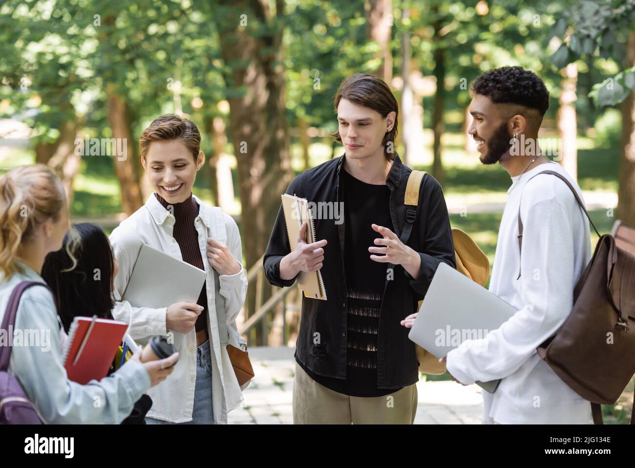 Studenti interrazziali positivi con computer portatili che parlano con amici sfocati nel parco Foto Stock