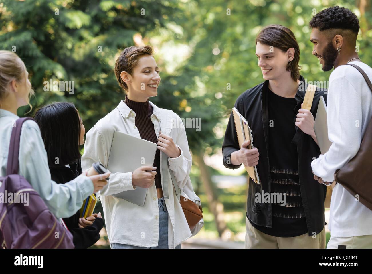 Gli studenti positivi che tengono notebook e parlano vicino ad amici interrazziali nel parco estivo Foto Stock