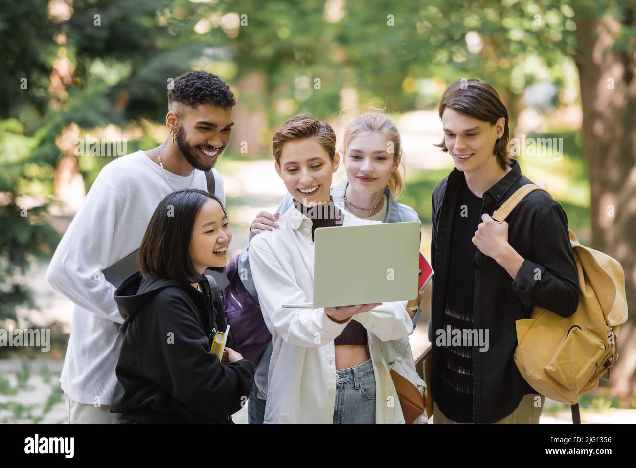 Sorridente studente che tiene un laptop vicino ad amici interrazziali nel parco estivo Foto Stock