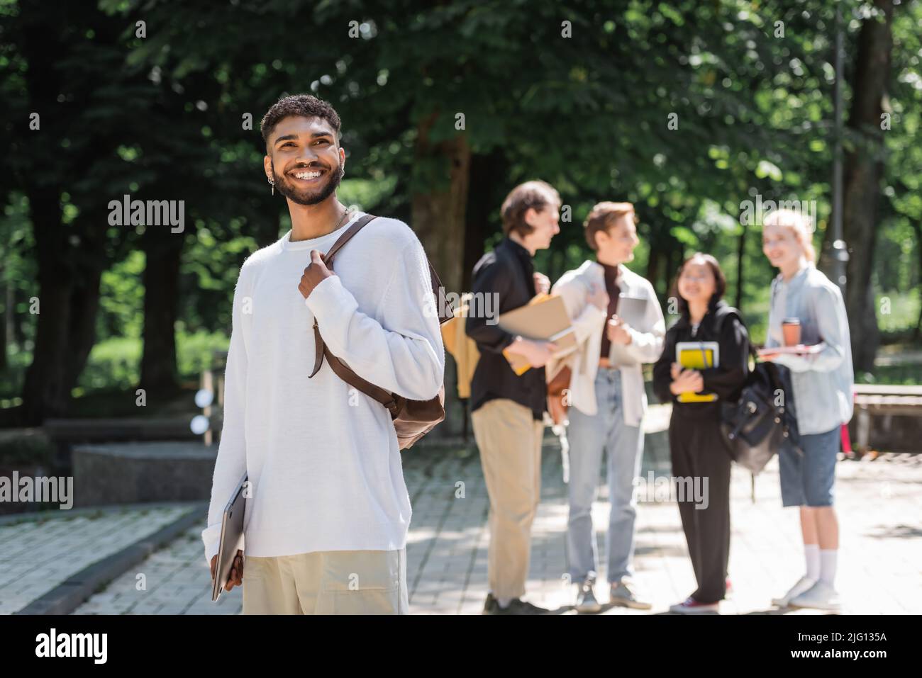 Sorridente studente afroamericano con un computer portatile in piedi vicino a amici sfocati nel parco estivo Foto Stock