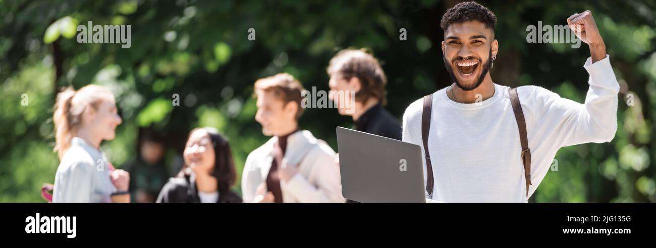 Eccitata studentessa afroamericana che tiene un laptop vicino a amici sfocati nel parco estivo, banner Foto Stock