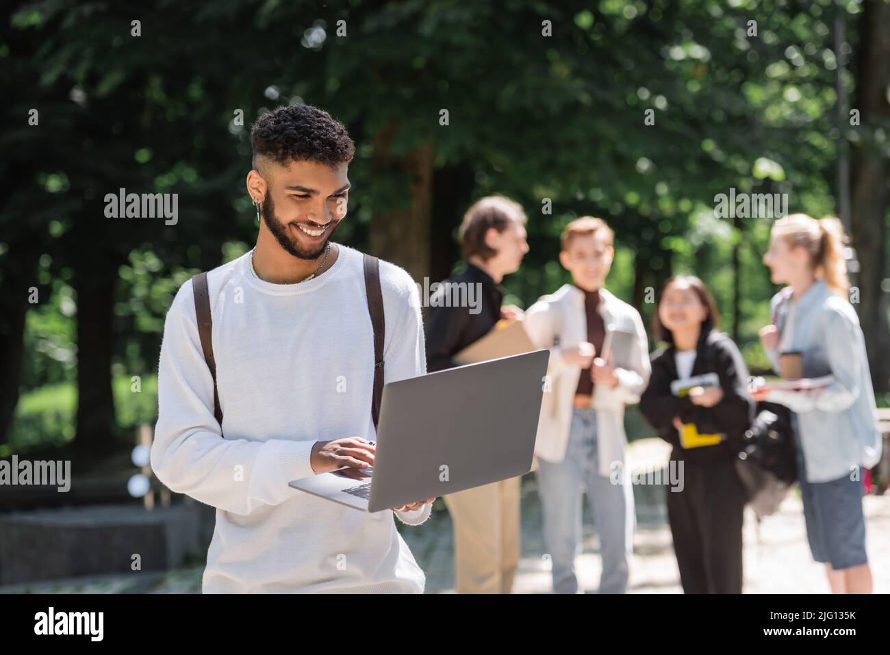 Sorridendo studente afroamericano usando il laptop vicino a blurred amici nel parco Foto Stock