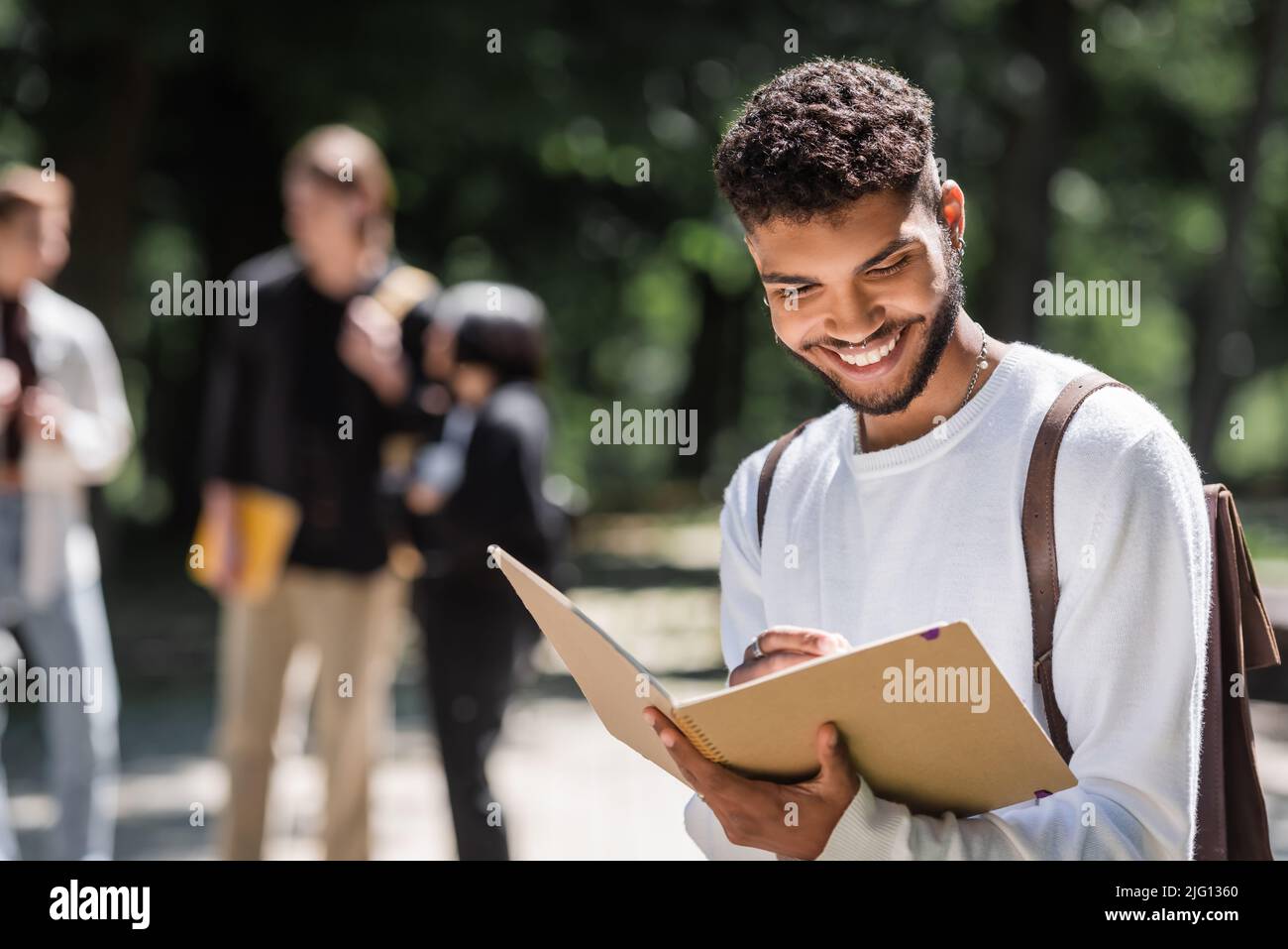 Sorridente studente afroamericano con scrittura zaino su notebook all'aperto Foto Stock
