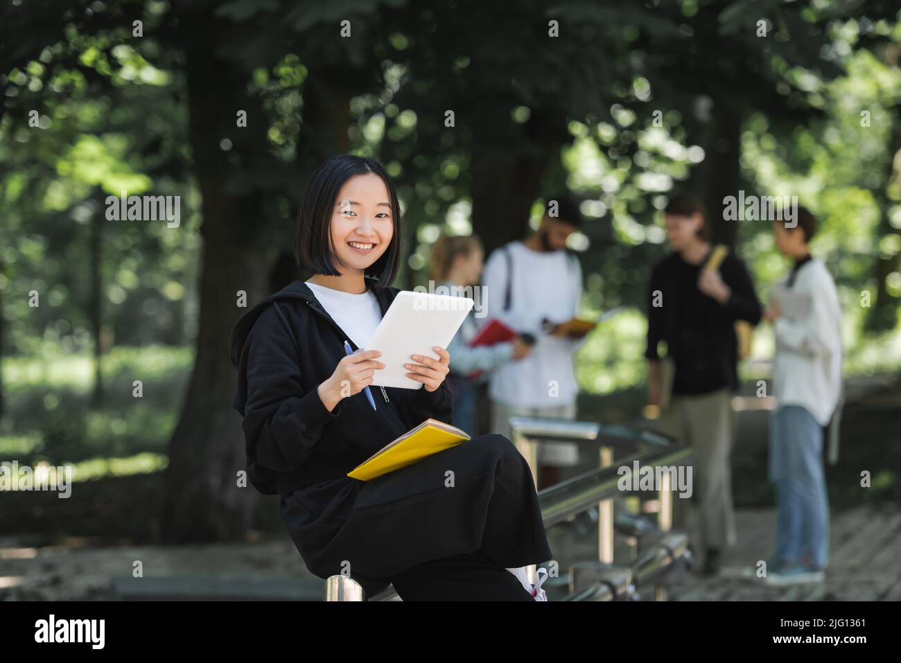 Allegro studente asiatico con tablet digitale e notebook guardando la macchina fotografica nel parco Foto Stock