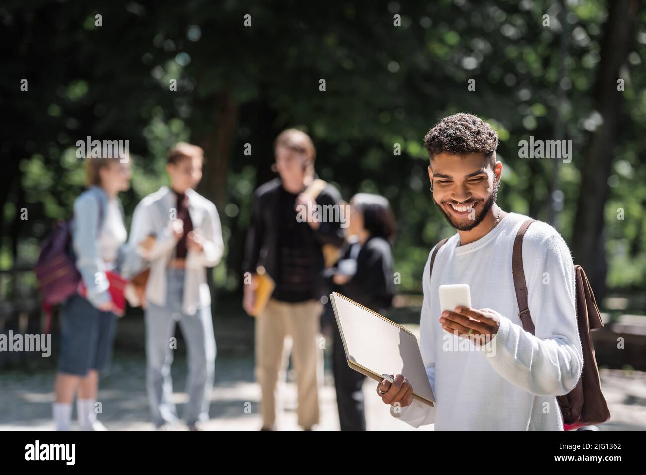 Sorridendo studente afroamericano che usa lo smartphone e tiene il notebook in parco Foto Stock