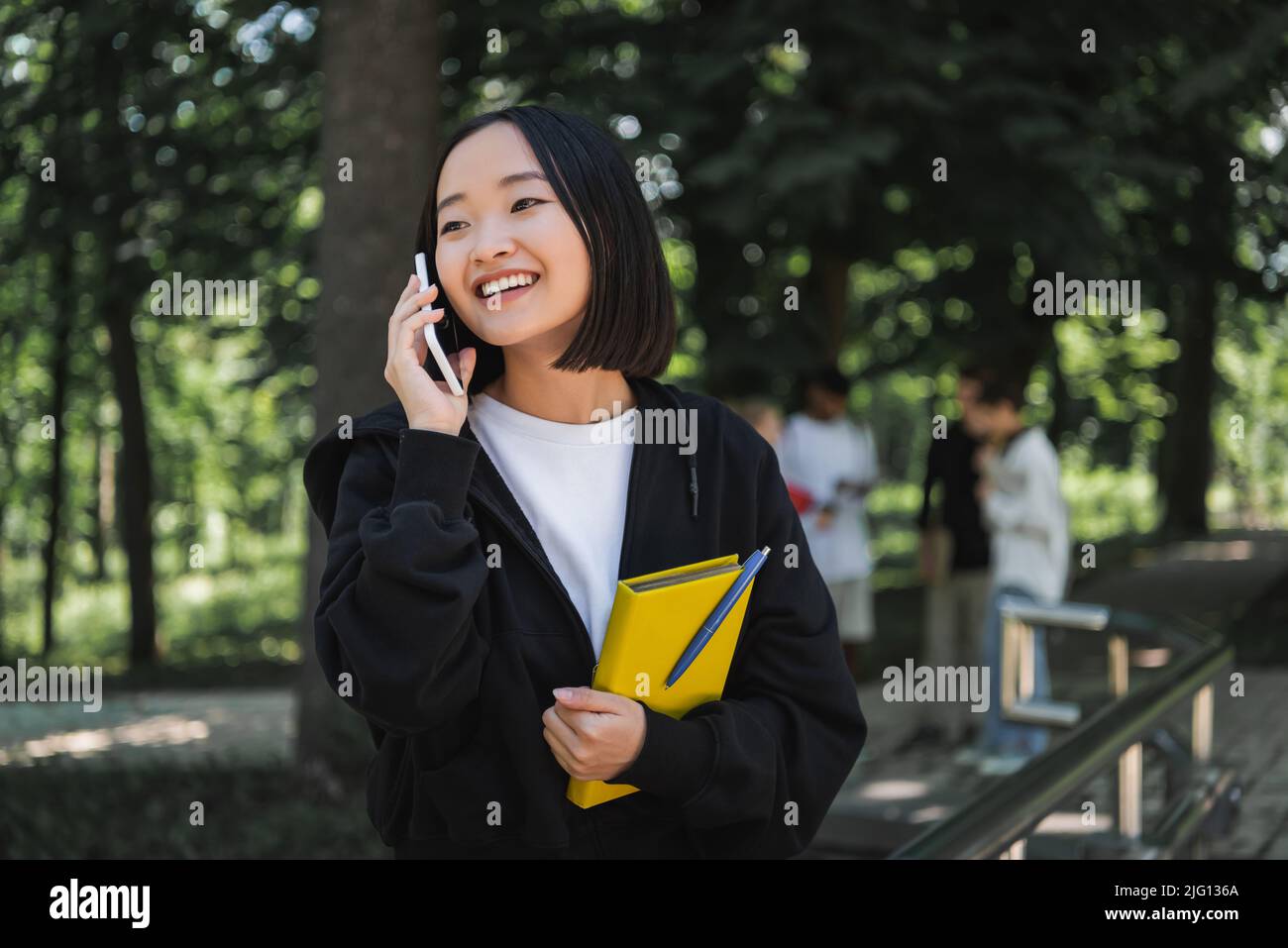 Positivo studente asiatico che tiene un libro e parla sullo smartphone nel parco Foto Stock