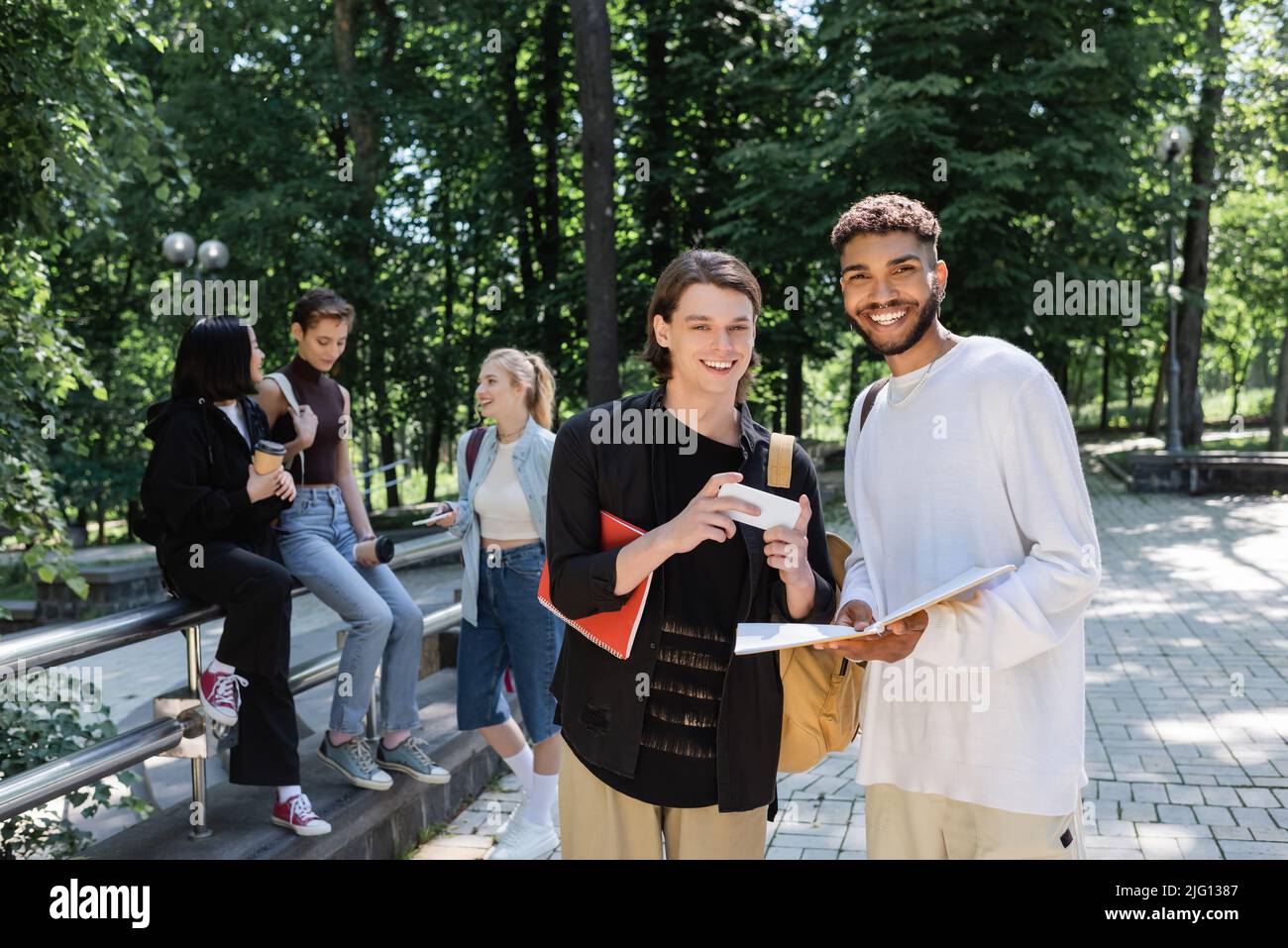 Studenti multietnici positivi con notebook e smartphone che osservano la telecamera nel parco Foto Stock