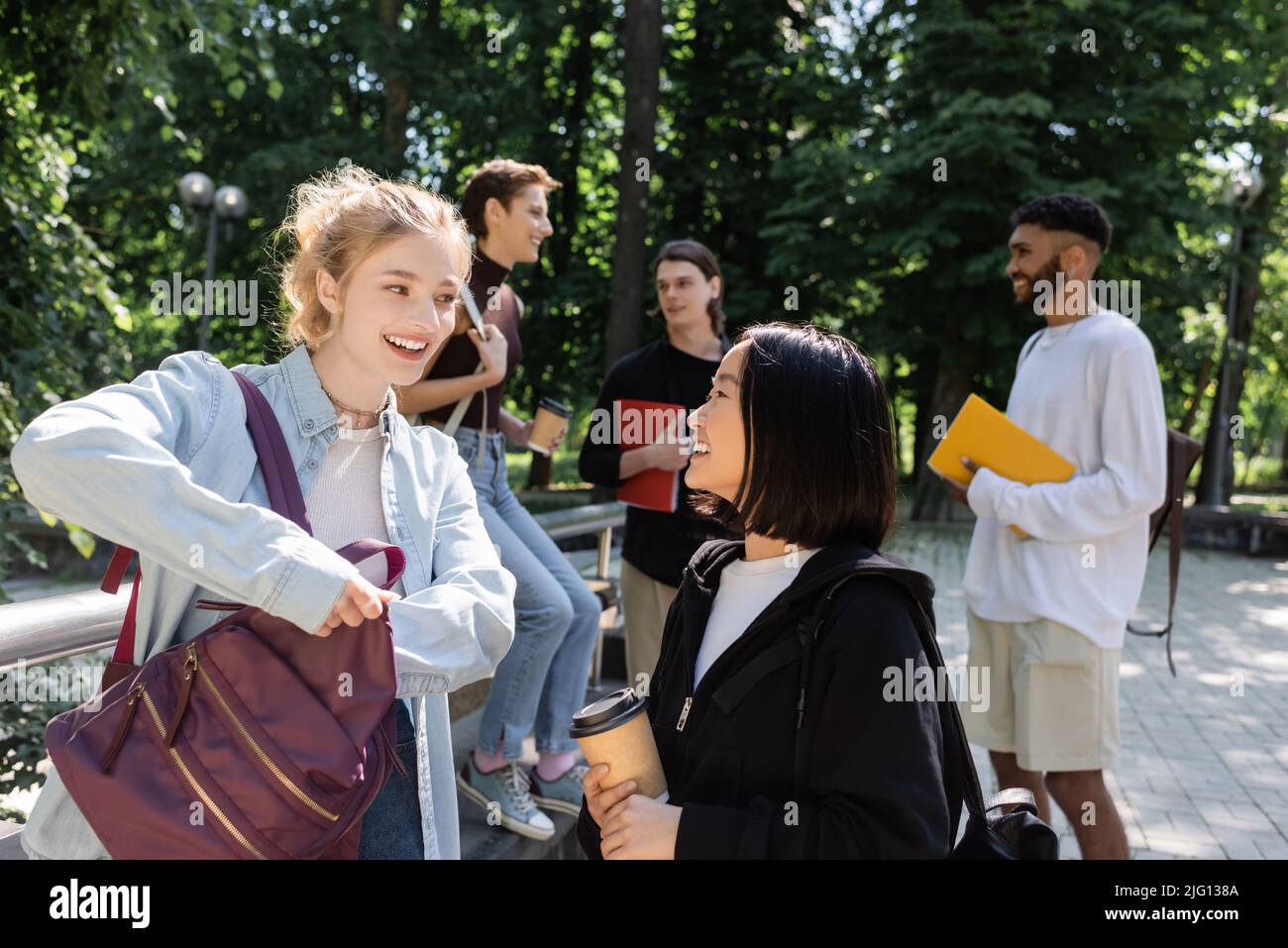 Studenti interrazziali positivi con caffè per andare a parlare vicino a blurred amici nel parco Foto Stock