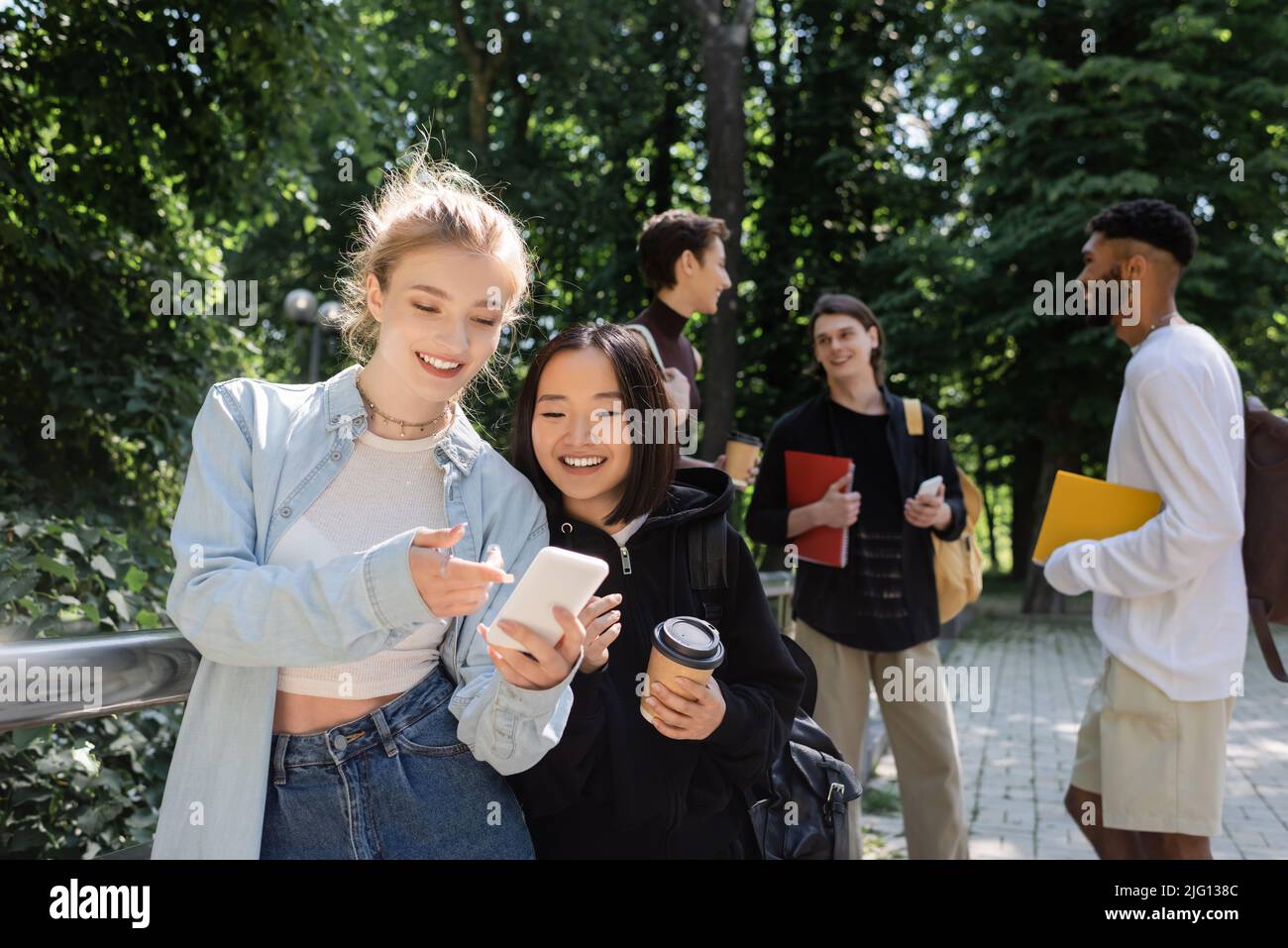 Sorridendo studente che punta con il dito sullo smartphone vicino a un amico asiatico con caffè per andare nel parco Foto Stock