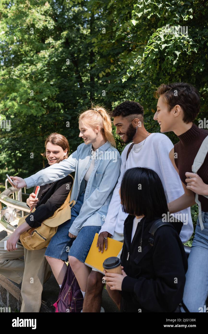 Sorridente studente che prende selfie su smartphone con amici multietnici nel parco Foto Stock