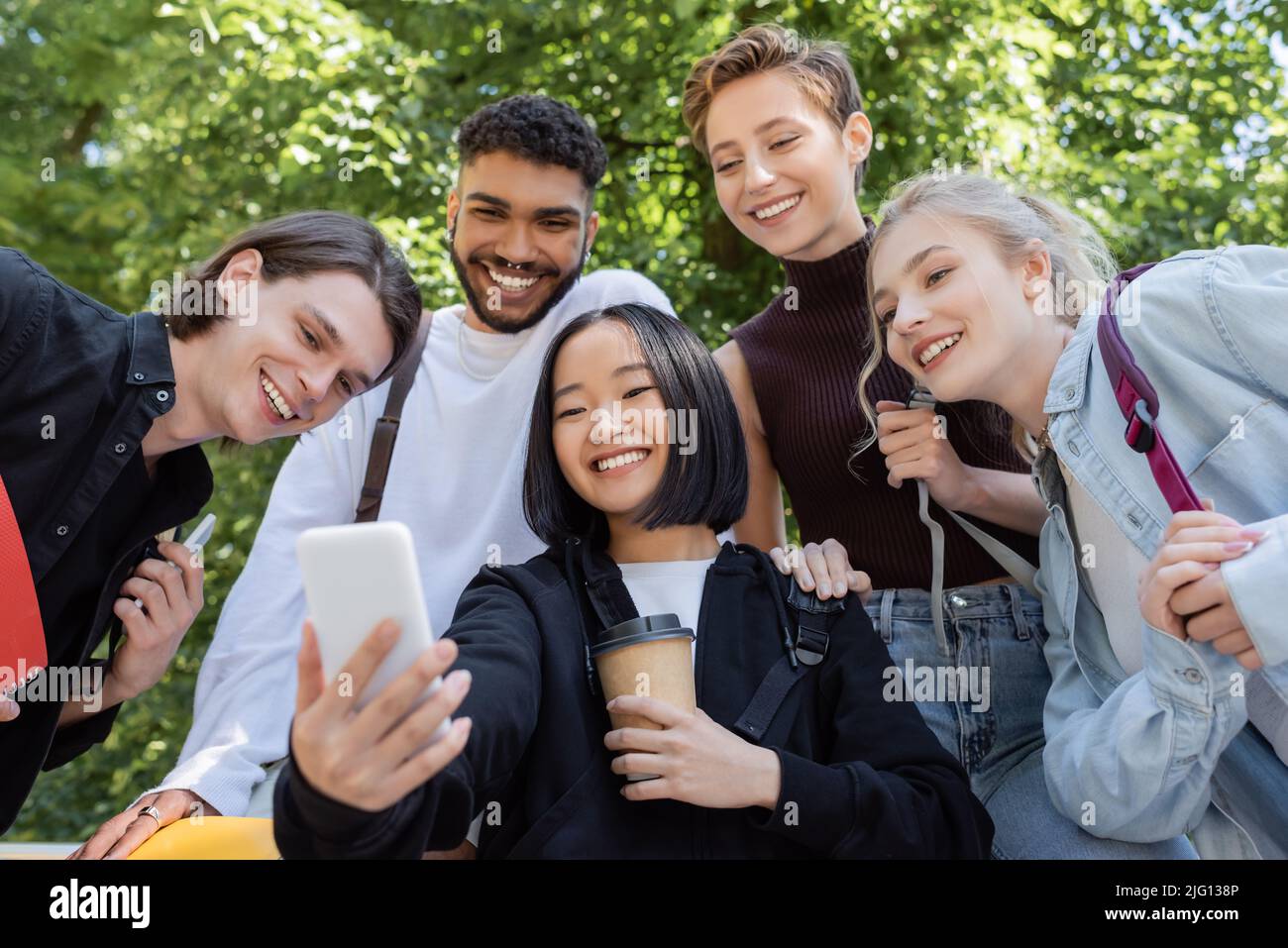 Studente asiatico che tiene il caffè per andare e prendere selfie con gli amici interrazziali nel parco Foto Stock