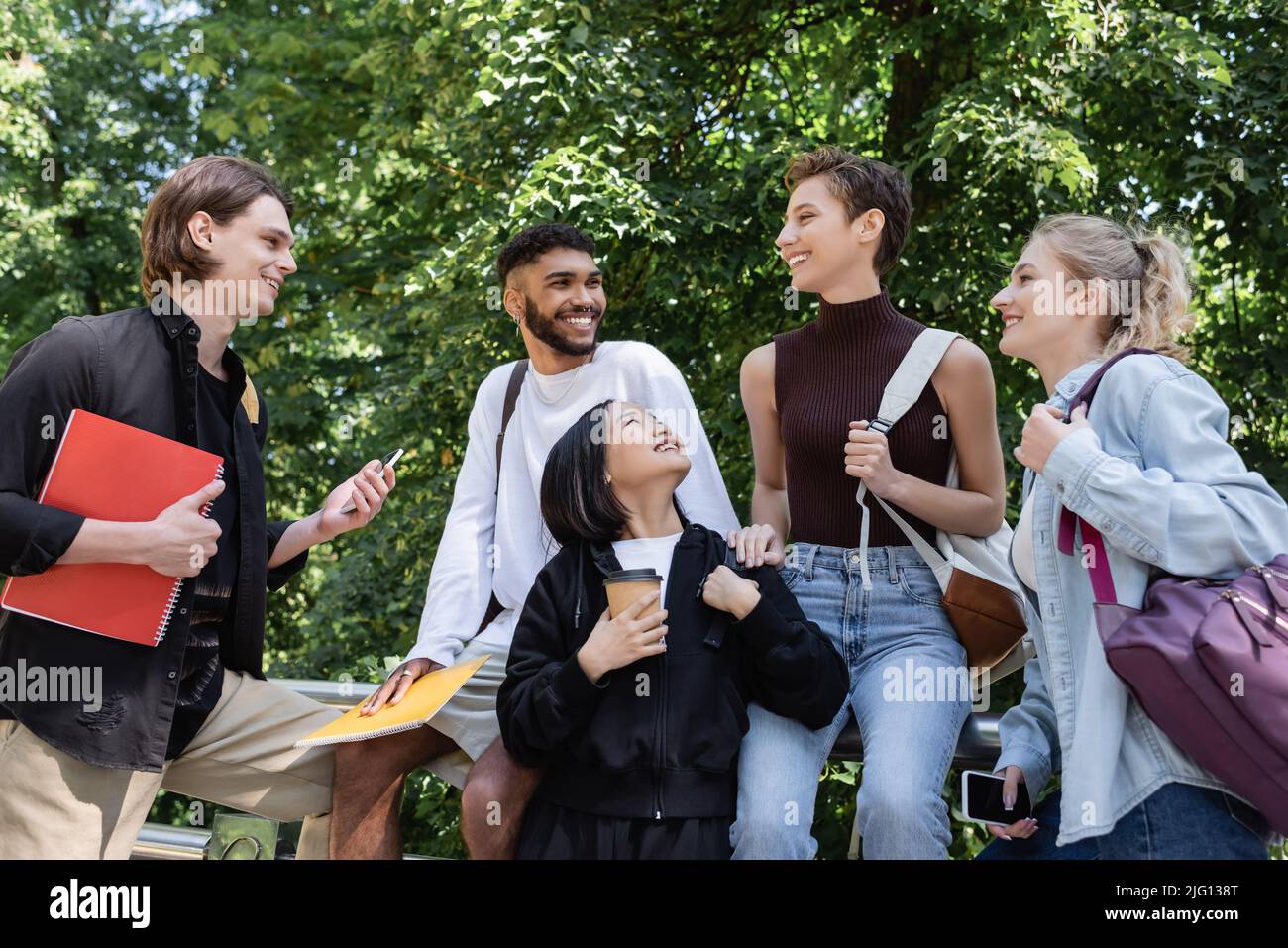 Sorridenti studenti multiculturali con notebook e smartphone che parlano in parco Foto Stock