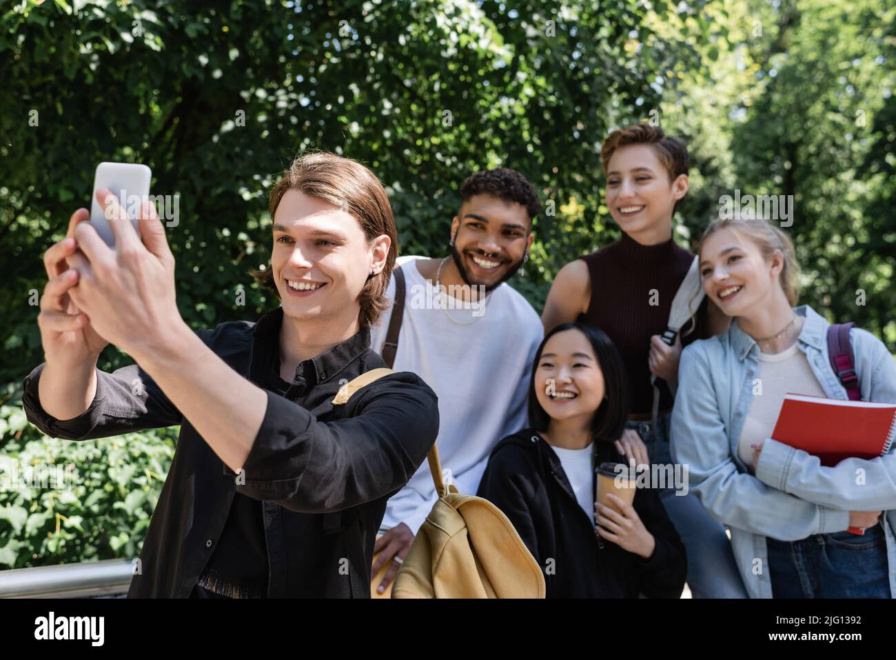 Sorridente studente che prende selfie su smartphone vicino ad amici interrazziali nel parco Foto Stock