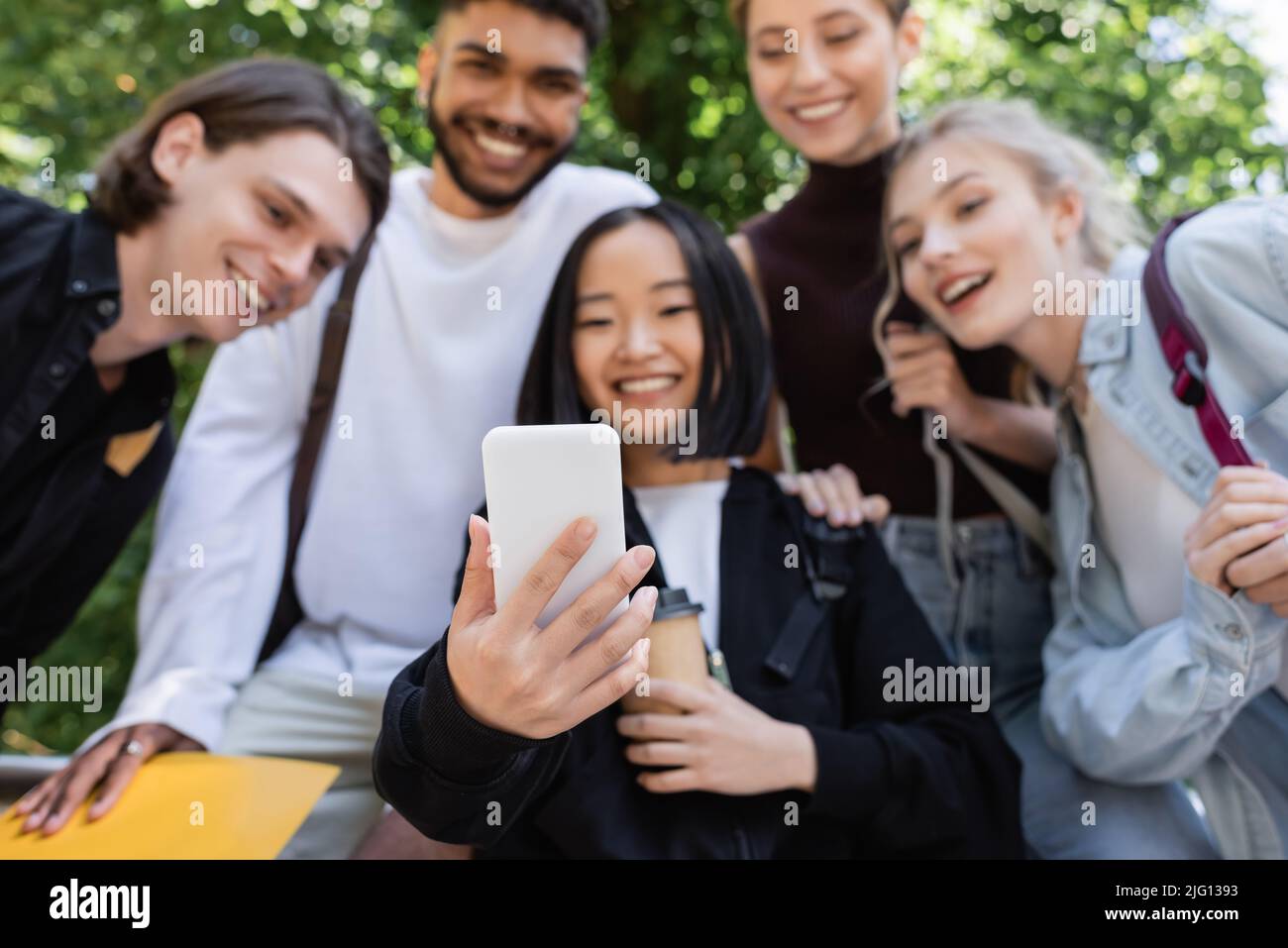 Offuscato studente asiatico tenendo smartphone vicino a amici interrazziali nel parco Foto Stock