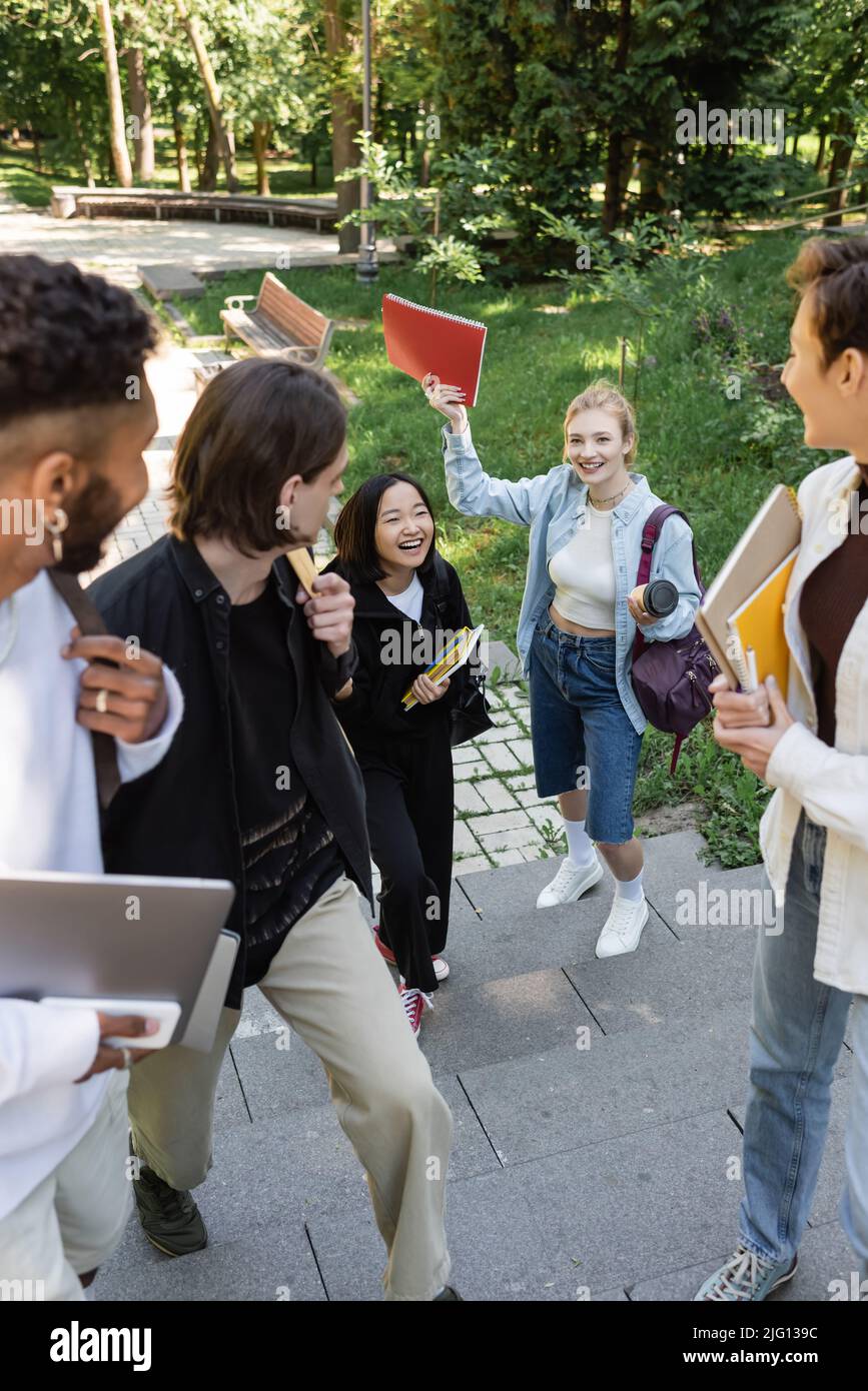 Studenti interrazziali con dispositivi che guardano amici allegri in parco Foto Stock