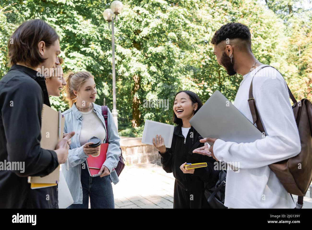 Studente asiatico che tiene un tablet digitale vicino a sorridenti amici multietnici nel parco estivo Foto Stock