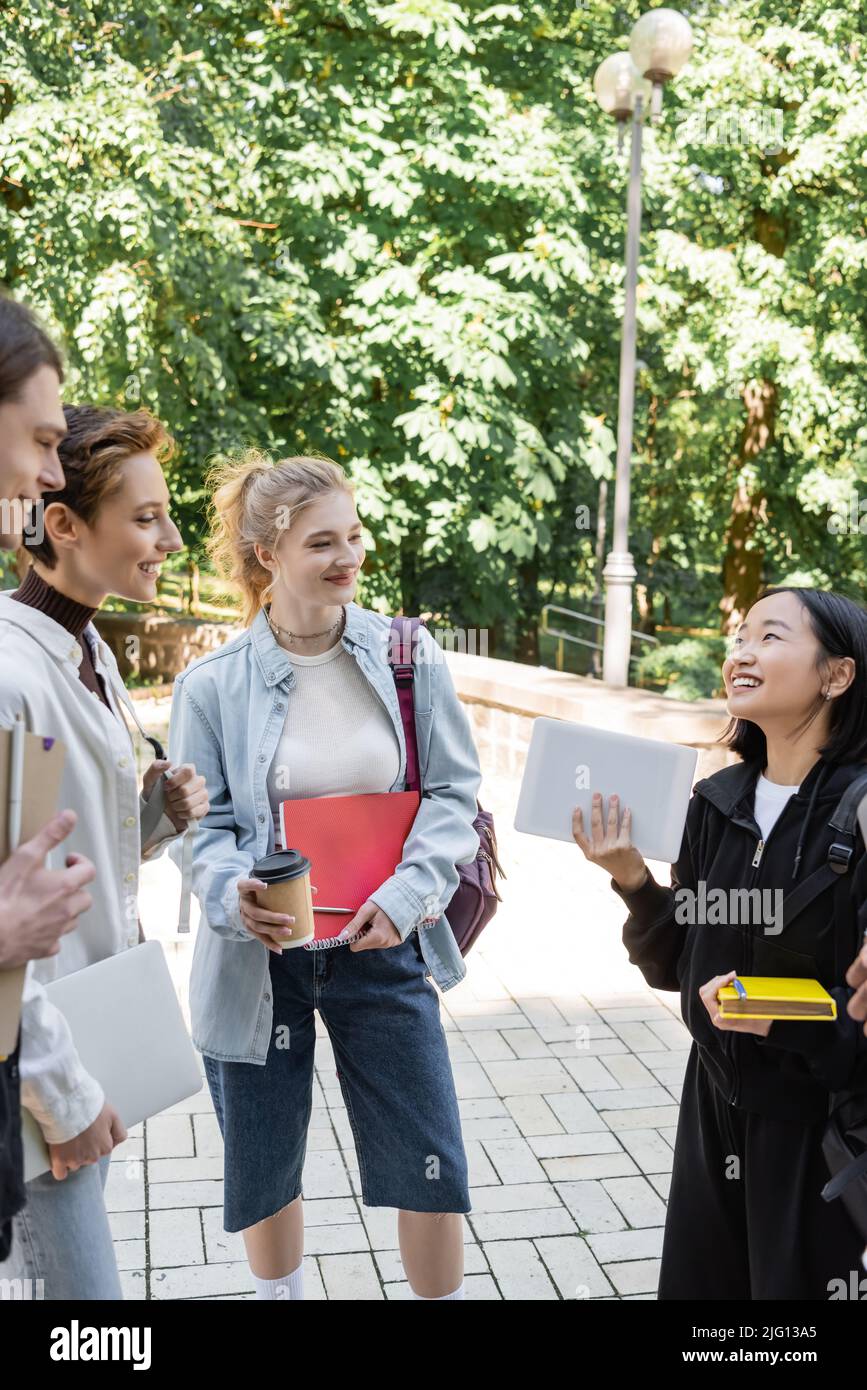 Sorridendo gli studenti con notebook che guardano un amico asiatico con un tablet digitale nel parco Foto Stock