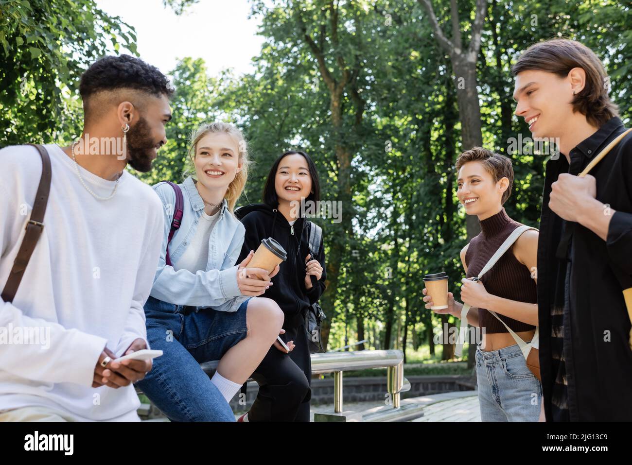 Positivo studente afroamericano utilizzando smartphone vicino ad amici interrazziali con caffè nel parco Foto Stock