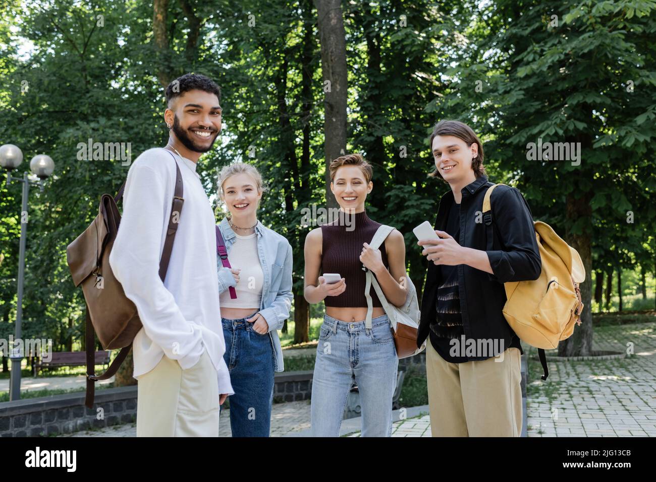 Sorridenti studenti interrazziali con zaini e smartphone che guardano la macchina fotografica nel parco Foto Stock