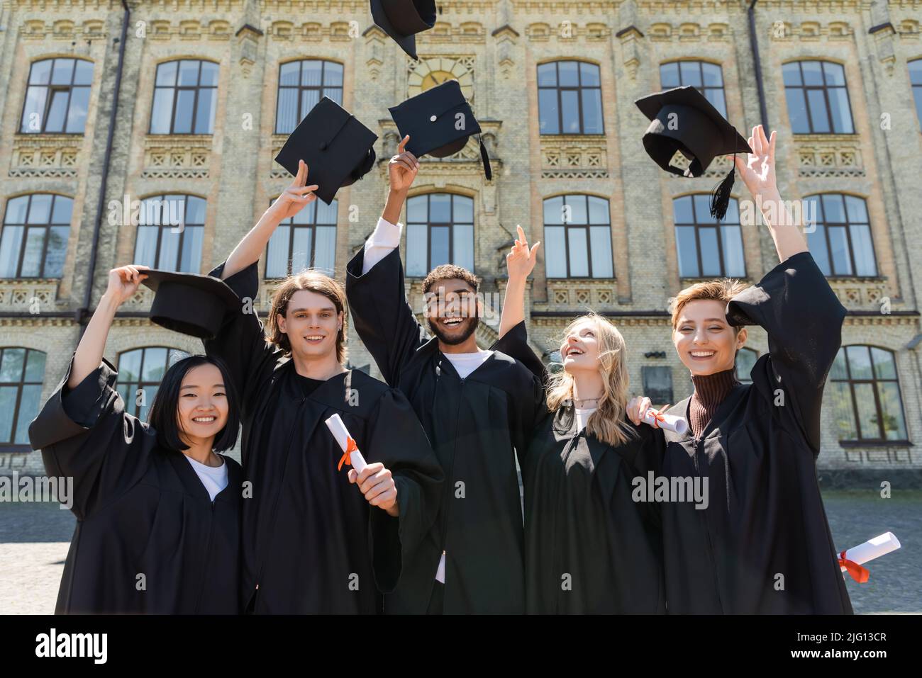 Sorridenti bachelor multietnici che tengono diplomi e lanciano cappellini all'aperto Foto Stock
