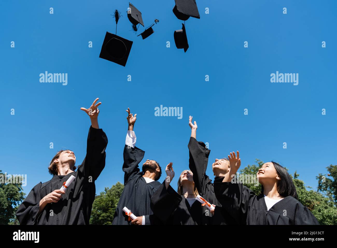 Bachelor interrazziali con diplomi che lanciano cappellini con cielo sullo sfondo Foto Stock