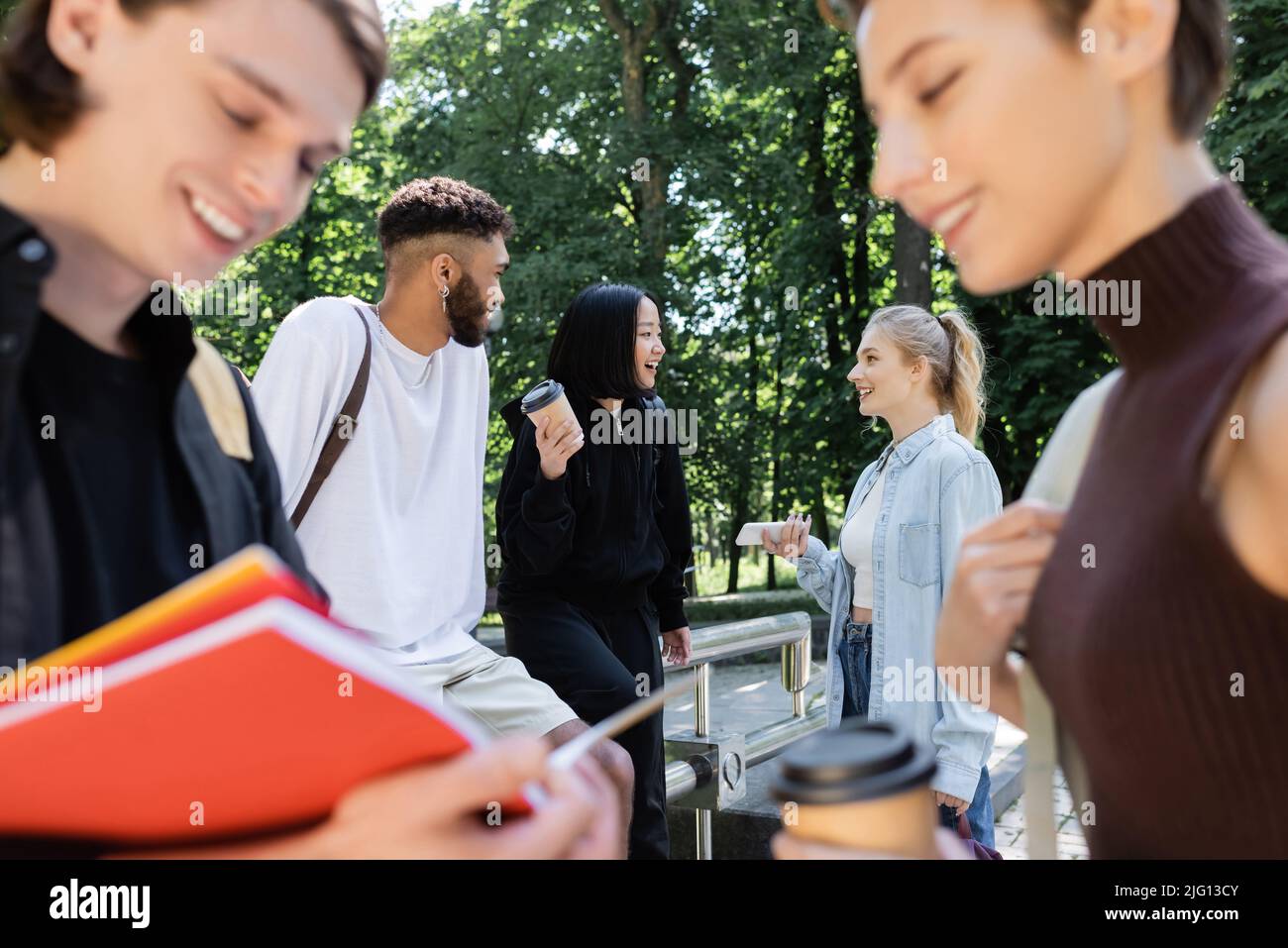 Sorridendo amici multietnici con caffè e smartphone che parlano vicino a amici sfocati nel parco Foto Stock