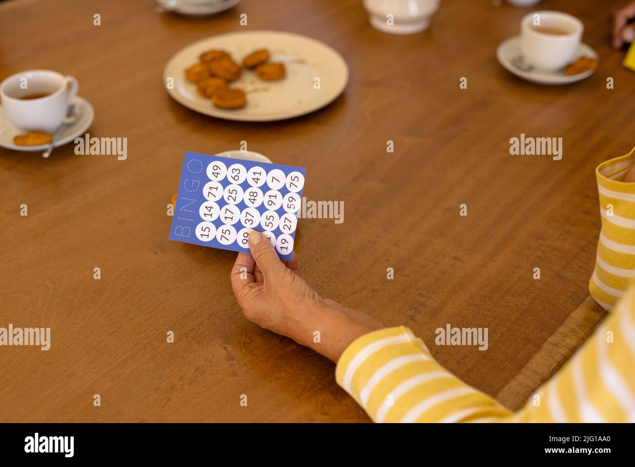 Mano ritagliata di donna anziana biraciale che tiene la scheda bingo al tavolo da pranzo in casa di cura Foto Stock