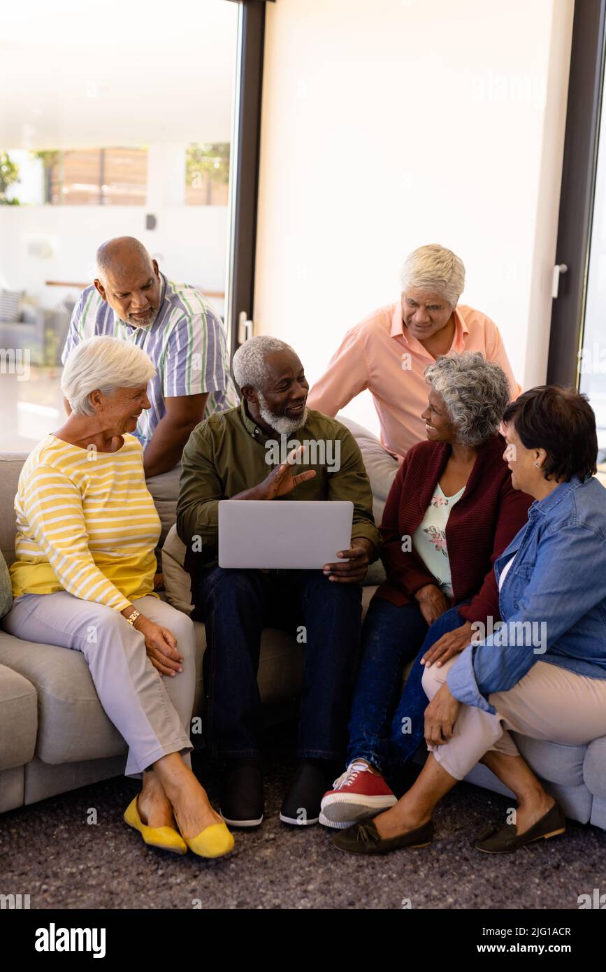 Uomo anziano multirazziale che tiene un laptop che parla con gli amici mentre si siede sul divano in casa di cura Foto Stock