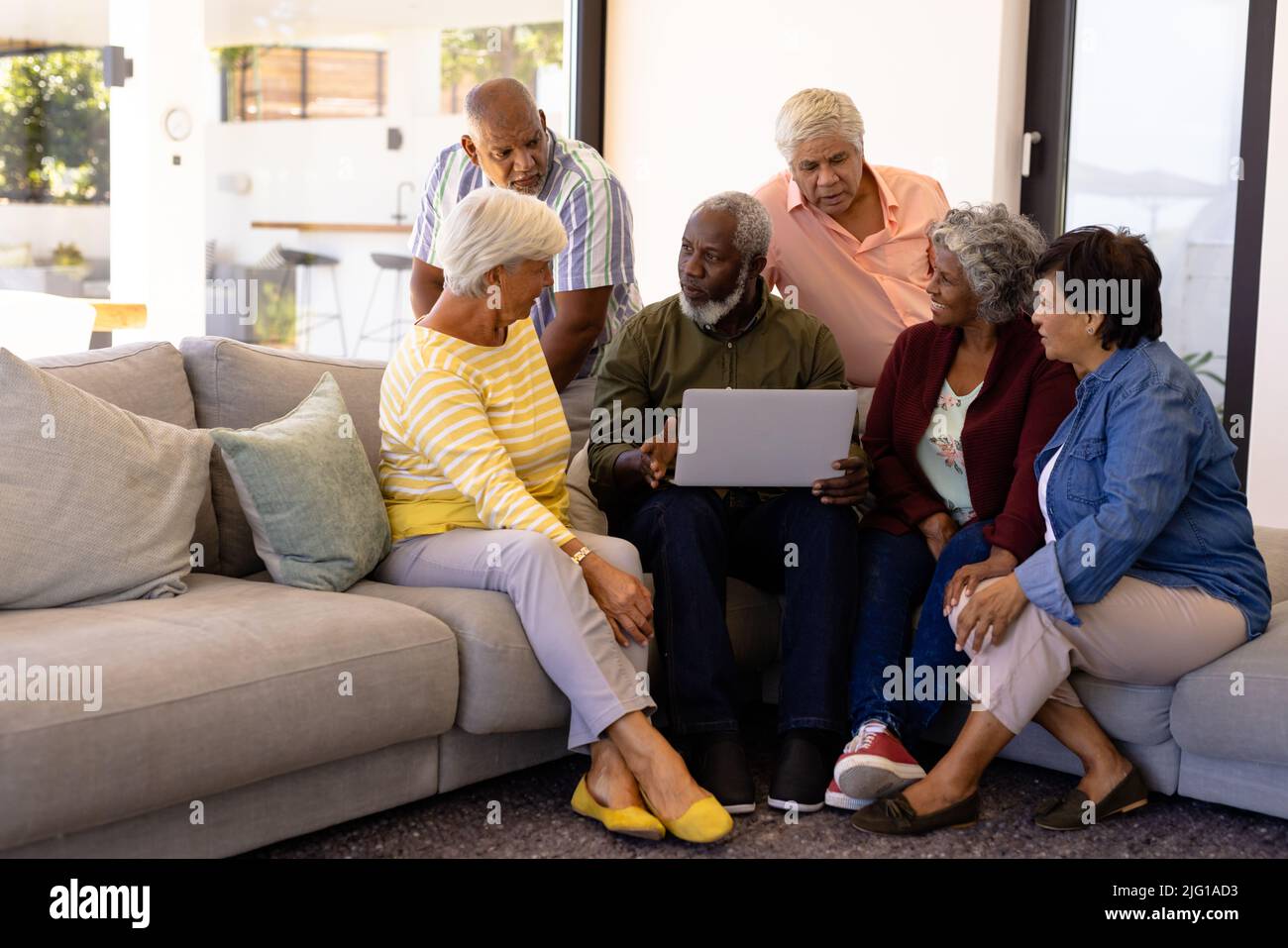 Uomo anziano multirazziale che tiene un laptop che spiega agli amici mentre si siede sul divano in casa di cura Foto Stock