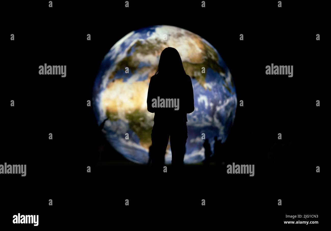 Silhouette di giovane donna in piedi di fronte al grande modello gonfiabile del pianeta Terra che risplende di notte, vista forma dietro Foto Stock