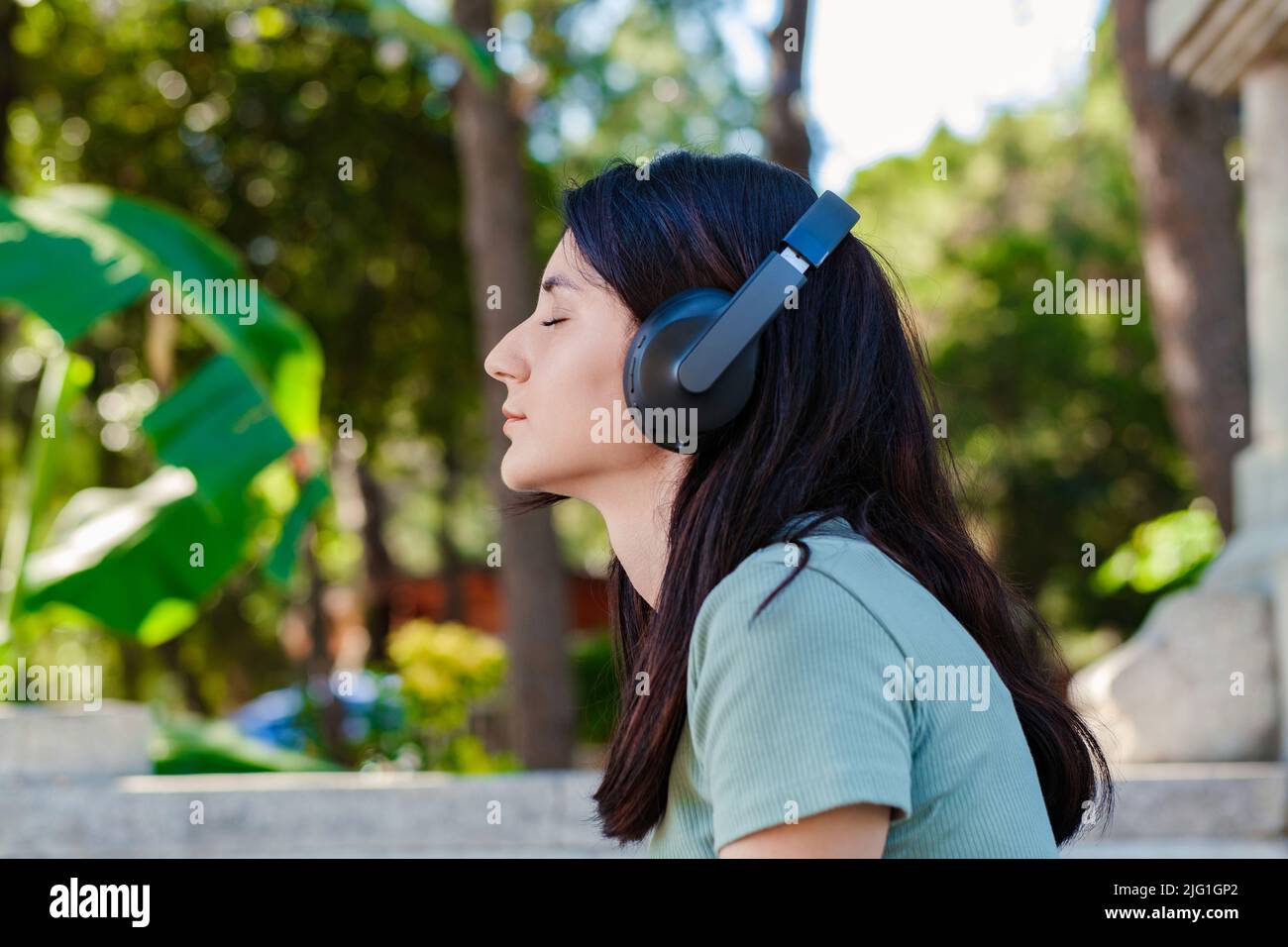 Vista laterale della donna millenaria carina con tee turchesi e cortine arancioni sul parco cittadino, all'aperto ascoltando musica con cuffie e occhi chiusi. Divergetevi Foto Stock