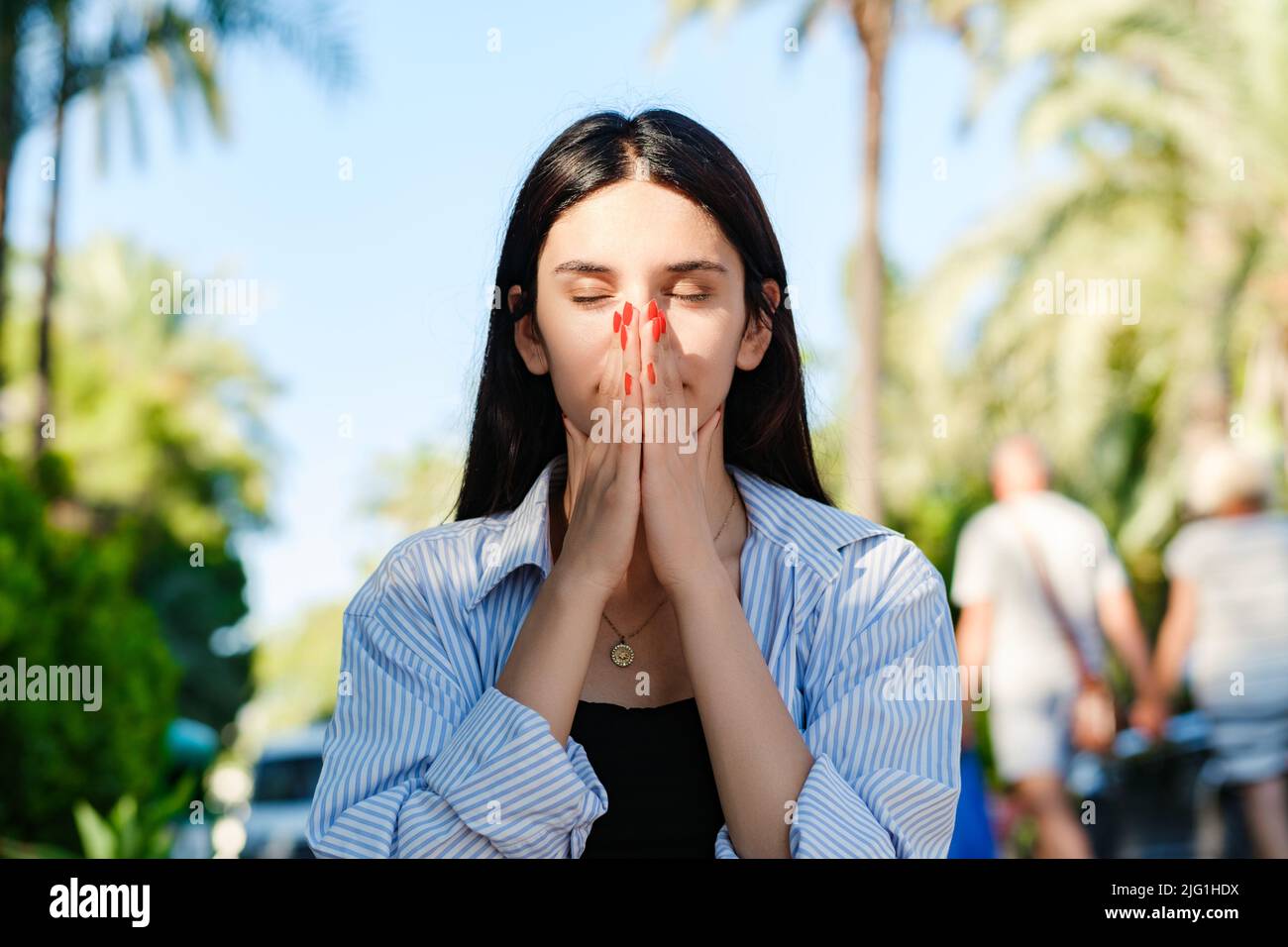 Ritratto di donna bruna carina con camicia a righe in piedi sul parco della città, esterno che copre il viso con le mani occhi chiusi sensazione di libertà e happine Foto Stock