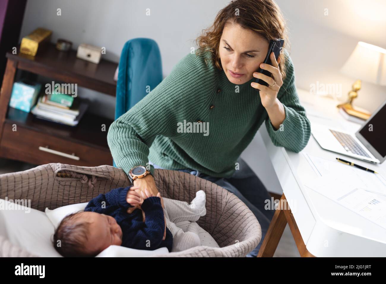 Madre caucasica di metà adulto che risponde allo smart phone mentre prende la cura del bambino neonato nella culla a casa Foto Stock